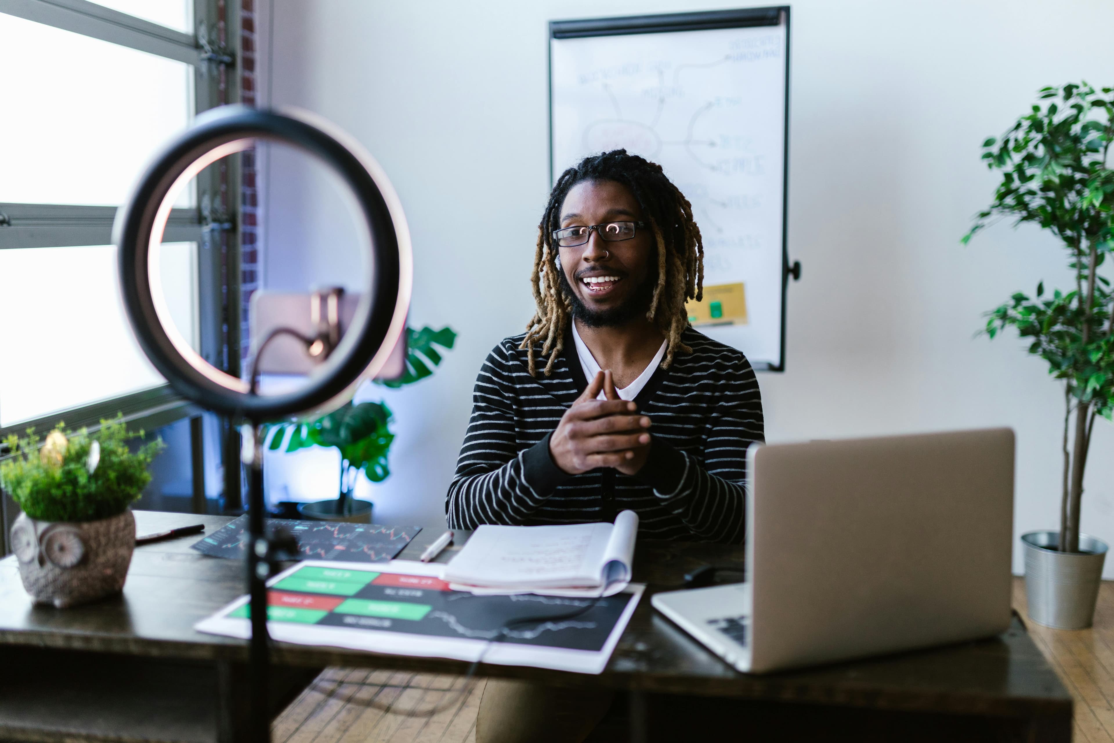 A man with dreadlocks sits at a desk, recording a video using a smartphone and a ring light setup, while explaining a concept on charts in the background. This setting answers "what is content marketing" by demonstrating how video and educational content is used to inform and build trust with an audience in professional industries.