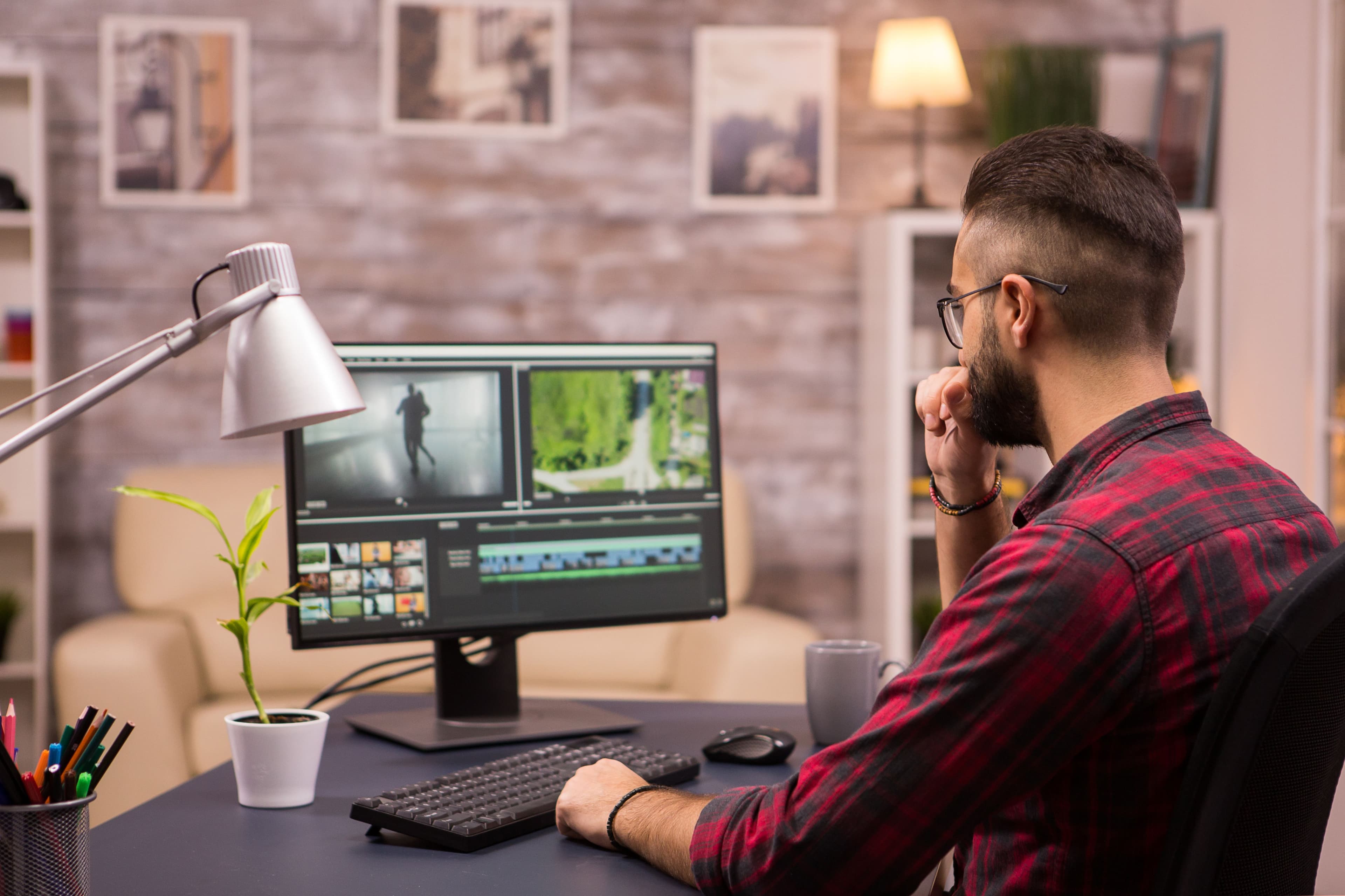 A man with glasses, wearing a red plaid shirt, is deeply focused on a computer screen with video editing software open, potentially taking a break to check the stock market performance today amidst his work.