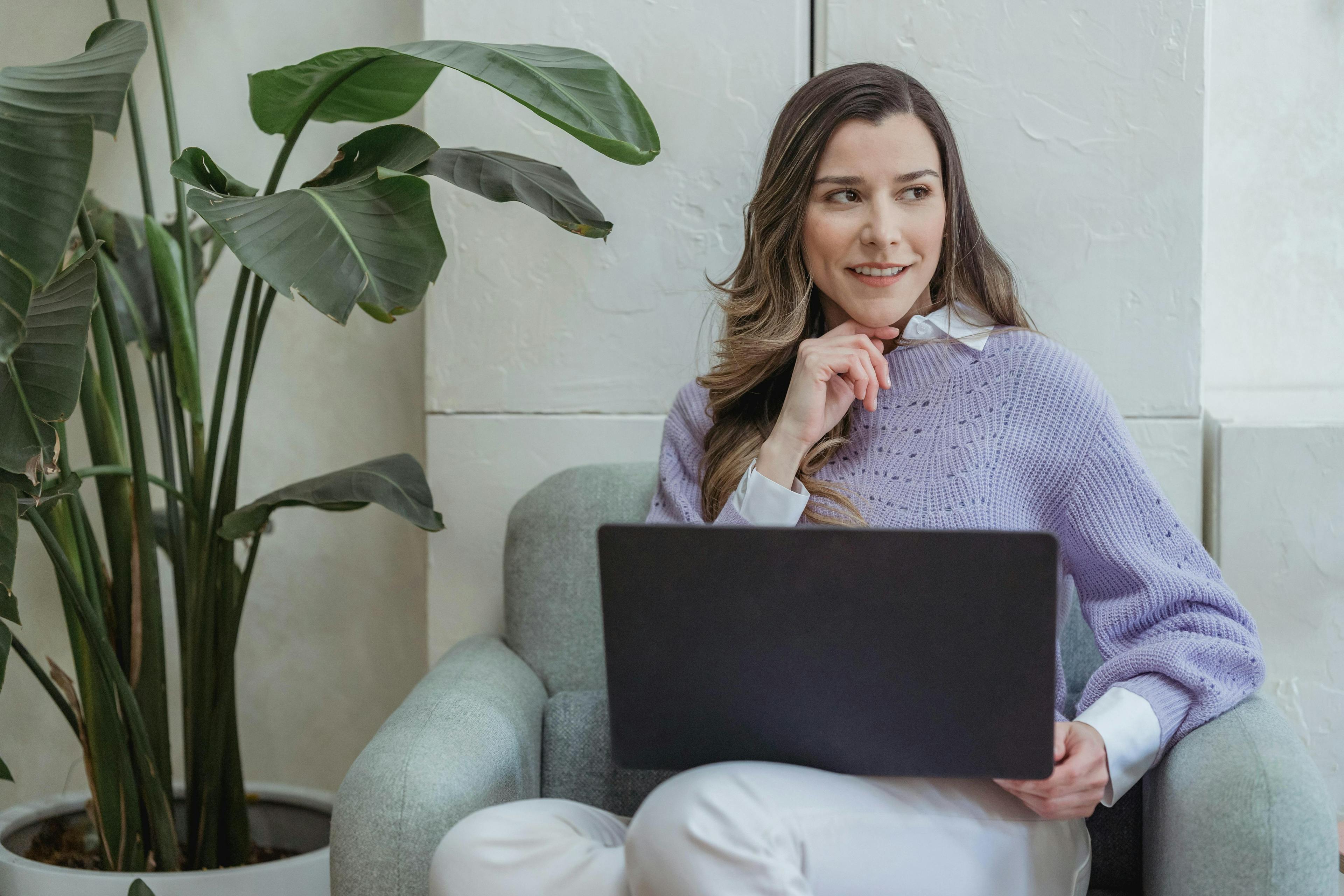 A woman sitting in an armchair with a laptop, smiling and thinking, with plants in the background. Inbound marketing funnel.