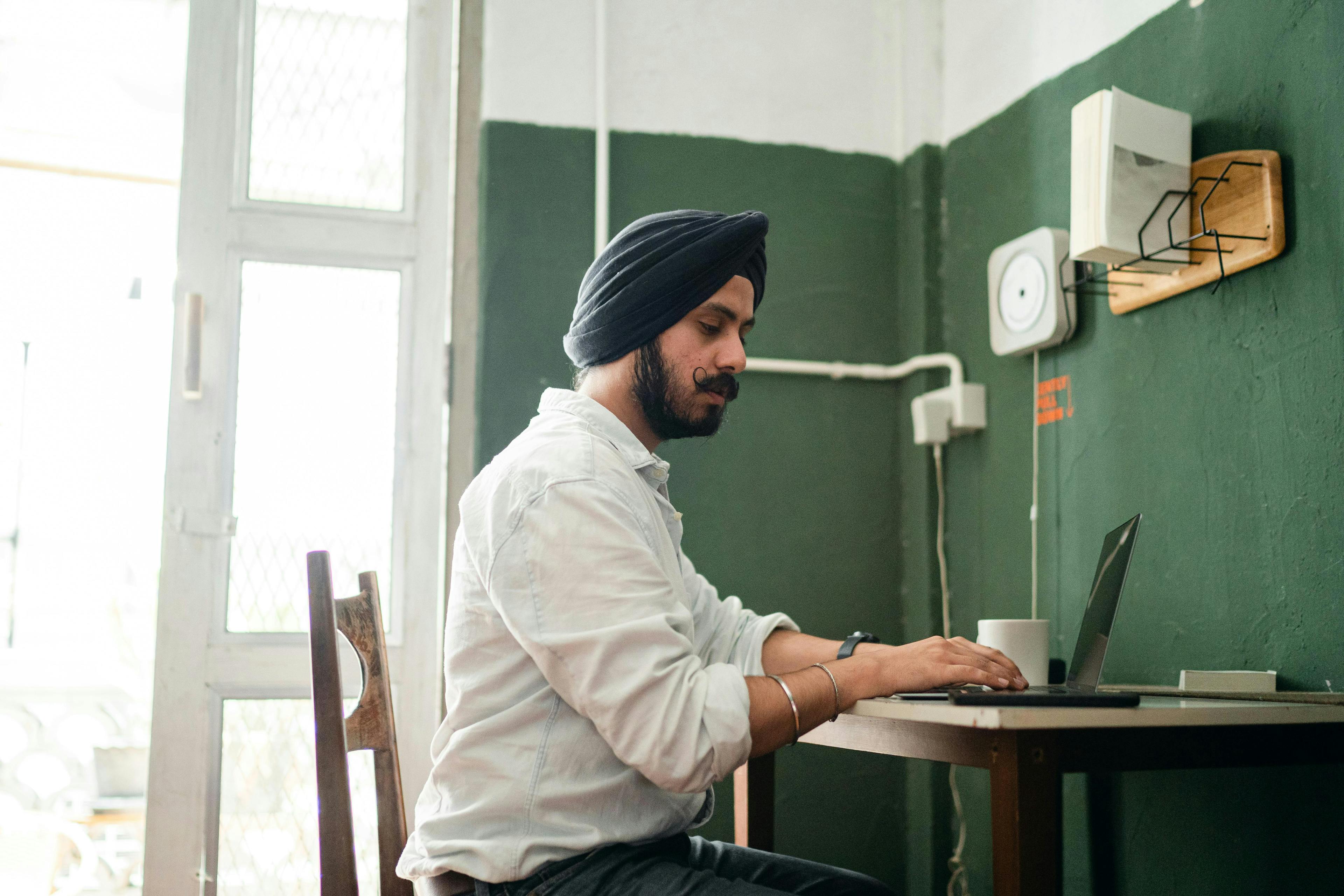 A man wearing a turban working on a laptop at a desk in a green-walled room. He appears focused, likely engaged in tasks related to marketing analytics jobs.