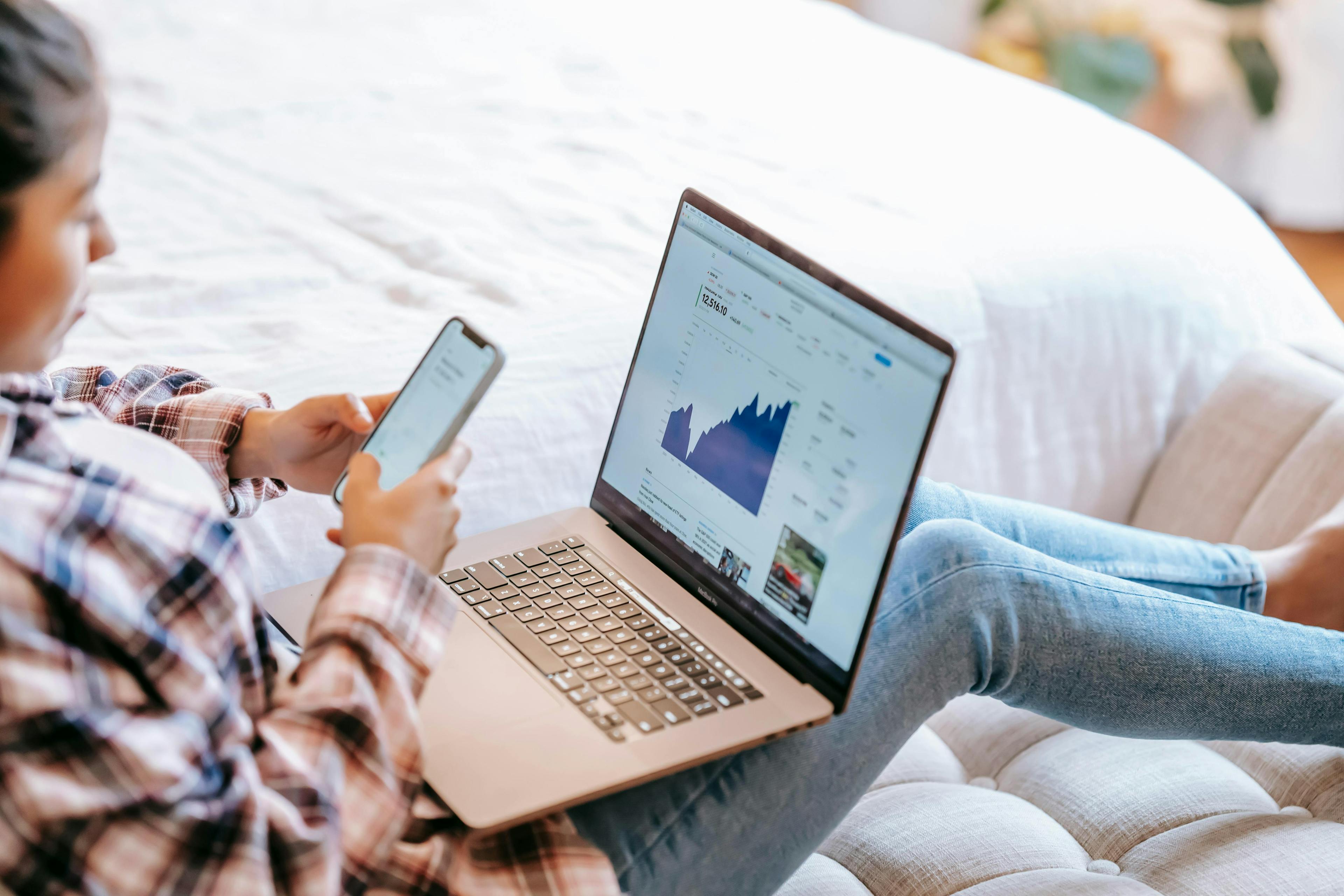 A person lounging on a bed is checking their phone while viewing graphs on their laptop screen, symbolizing influencer marketing services.