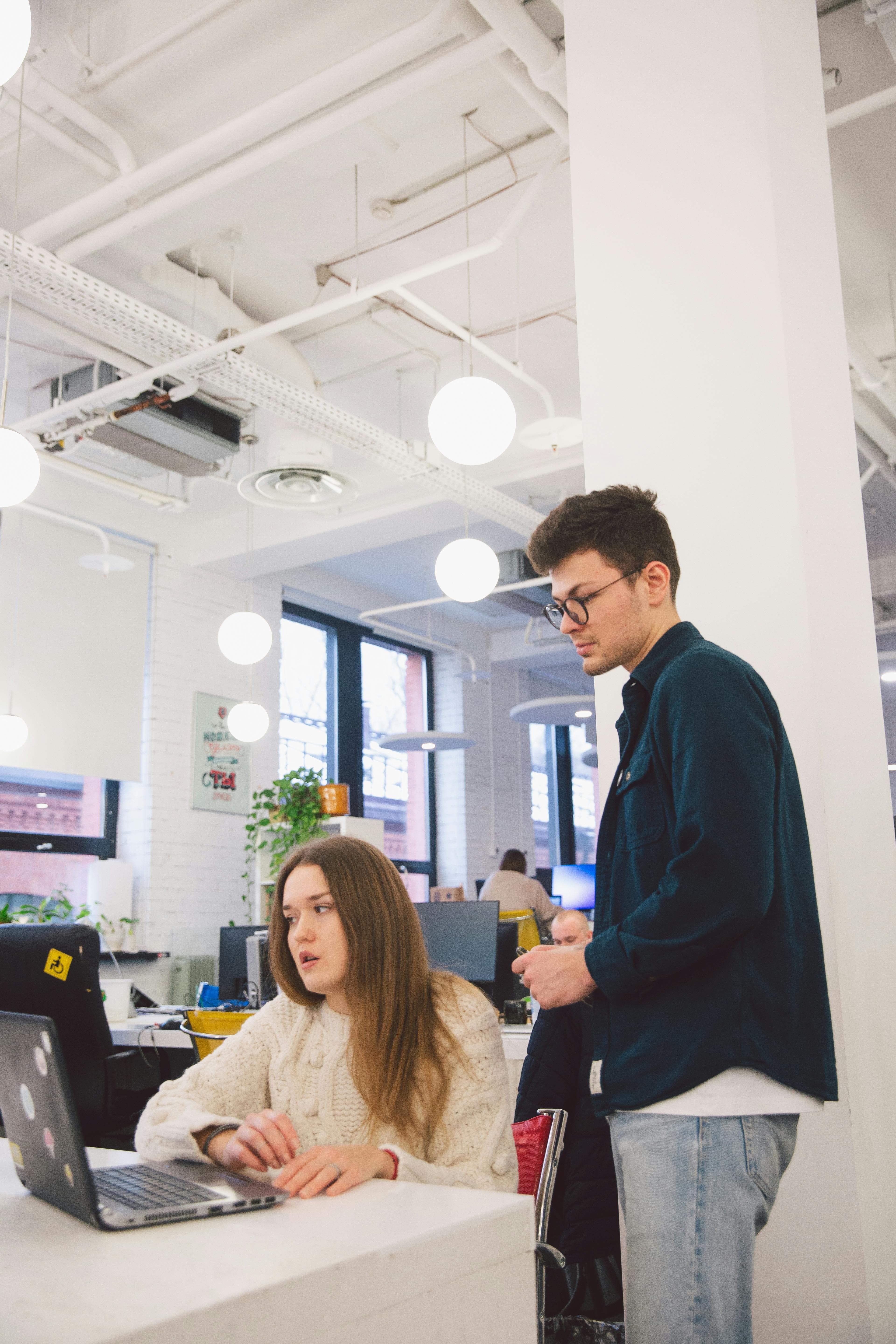 A collaborative office environment with a man standing and a woman sitting in front of a laptop, discussing a project. They might be going over the results of a split testing campaign, reviewing the data to see which version of their product or marketing strategy performed better.