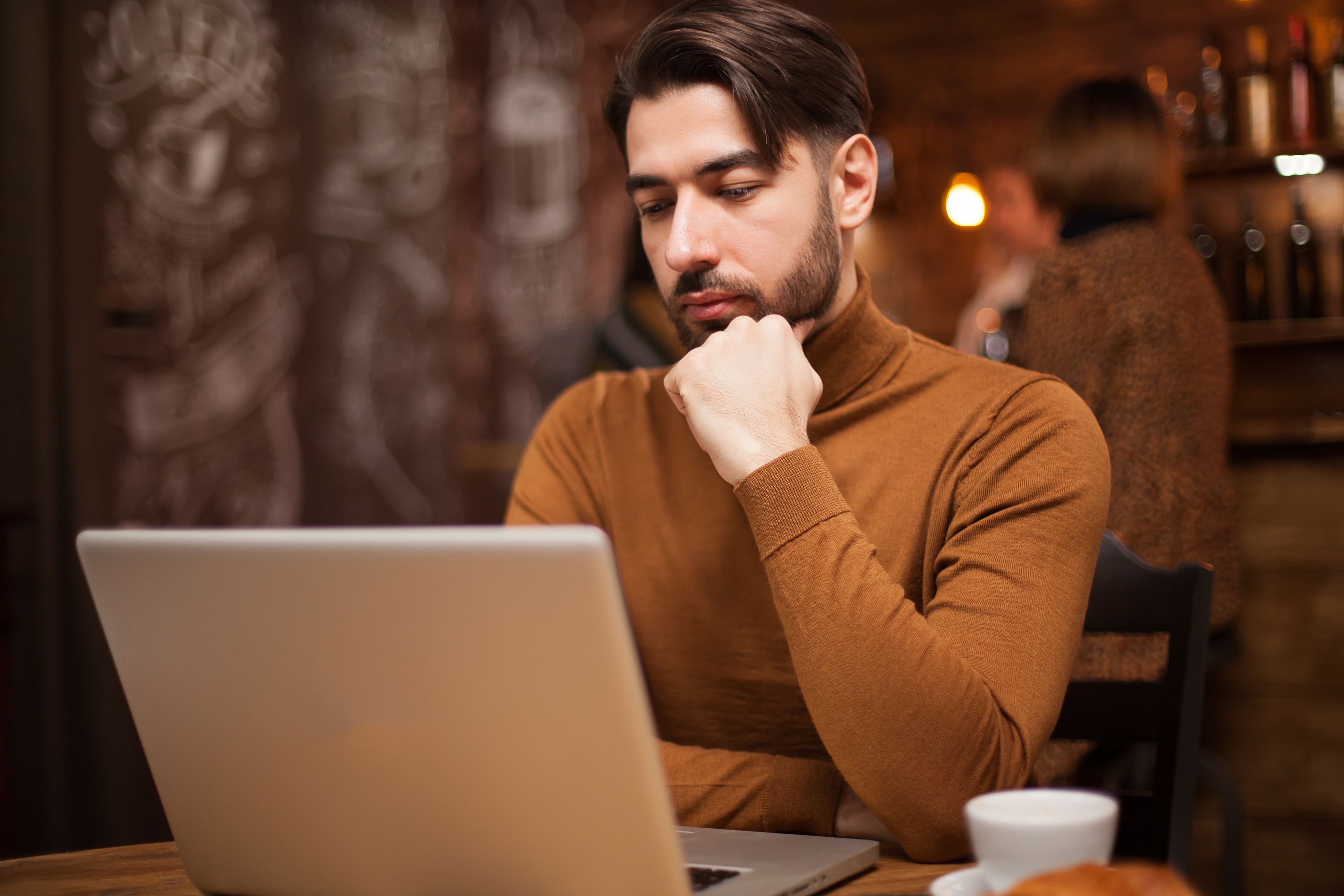 A man thoughtfully looking at his laptop screen while working in a coffee shop. The casual setting and his focused expression illustrate "what is DTC marketing" by emphasizing the importance of digital platforms for businesses directly marketing products to consumers without intermediaries.