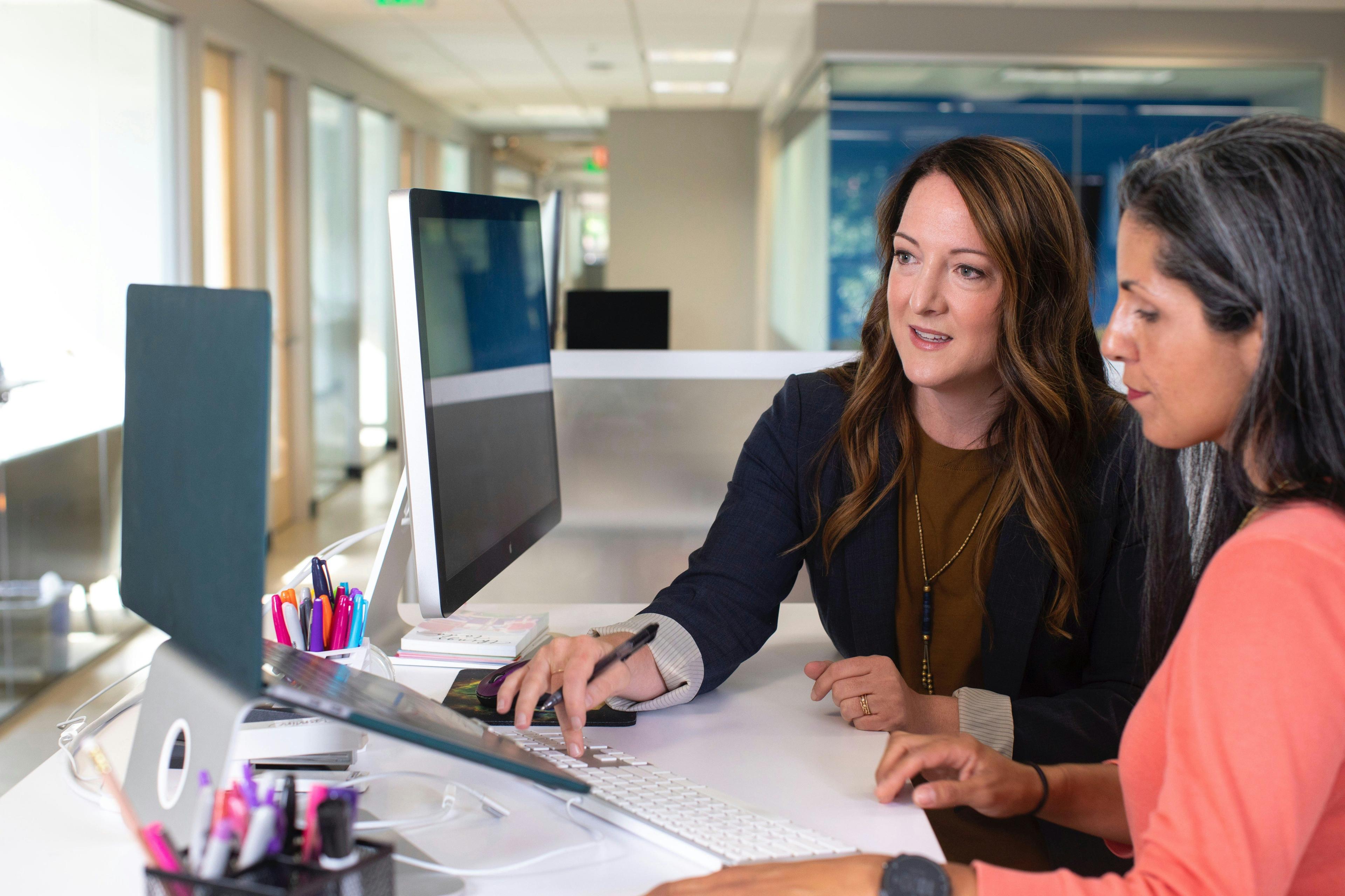 Two women working together at a computer desk in a modern office setting, discussing strategies for improving video content.