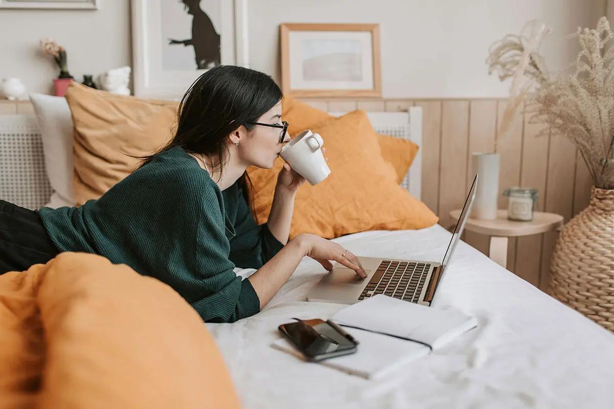 A woman is comfortably lying on a bed with orange pillows, wearing a dark green sweater and glasses. She is sipping from a white mug while using a laptop. Next to her on the bed are a smartphone and an open notebook. The cozy and relaxed setting is enhanced by framed art on the wall and a wicker basket on the side. This image can be associated with the phrase "black market performance."