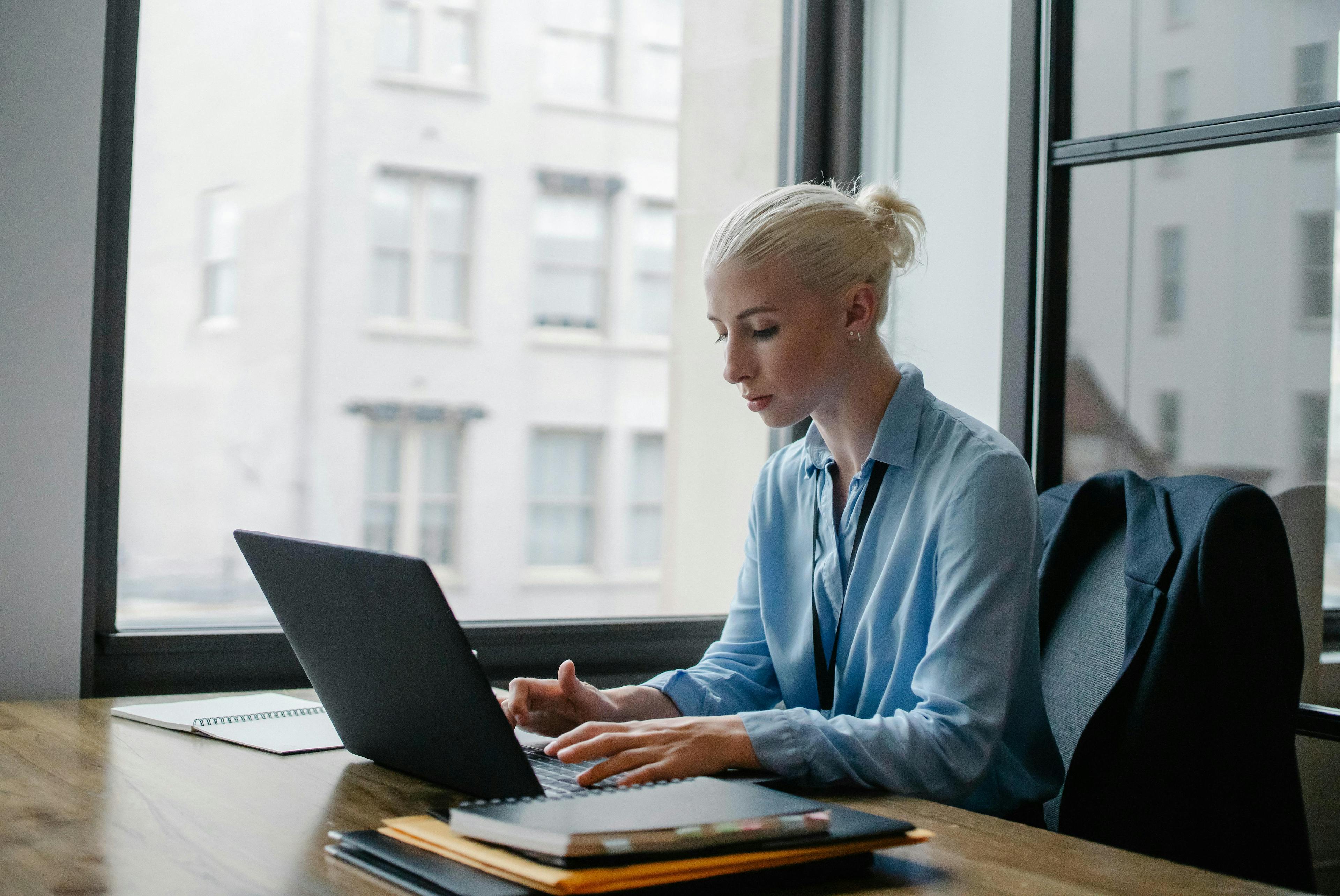 A focused businesswoman working on her laptop at a desk with a window view of city buildings, ideal for illustrating Shopify landing pages. The setting highlights professionalism and productivity, suitable for ecommerce and online business contexts.