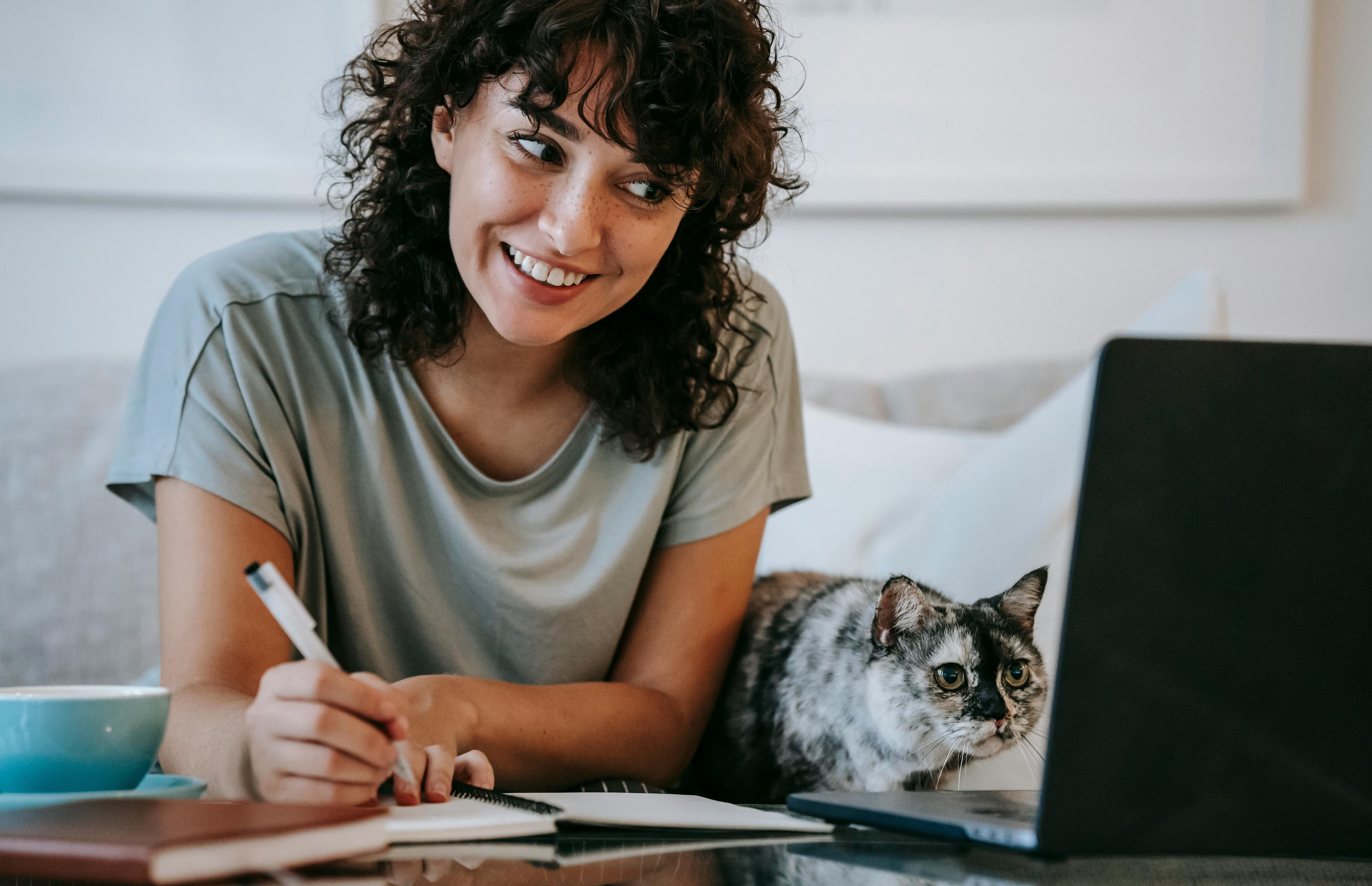 A woman smiling and writing notes while working on a laptop at home, accompanied by her cat, representing a digital marketing agency for small business