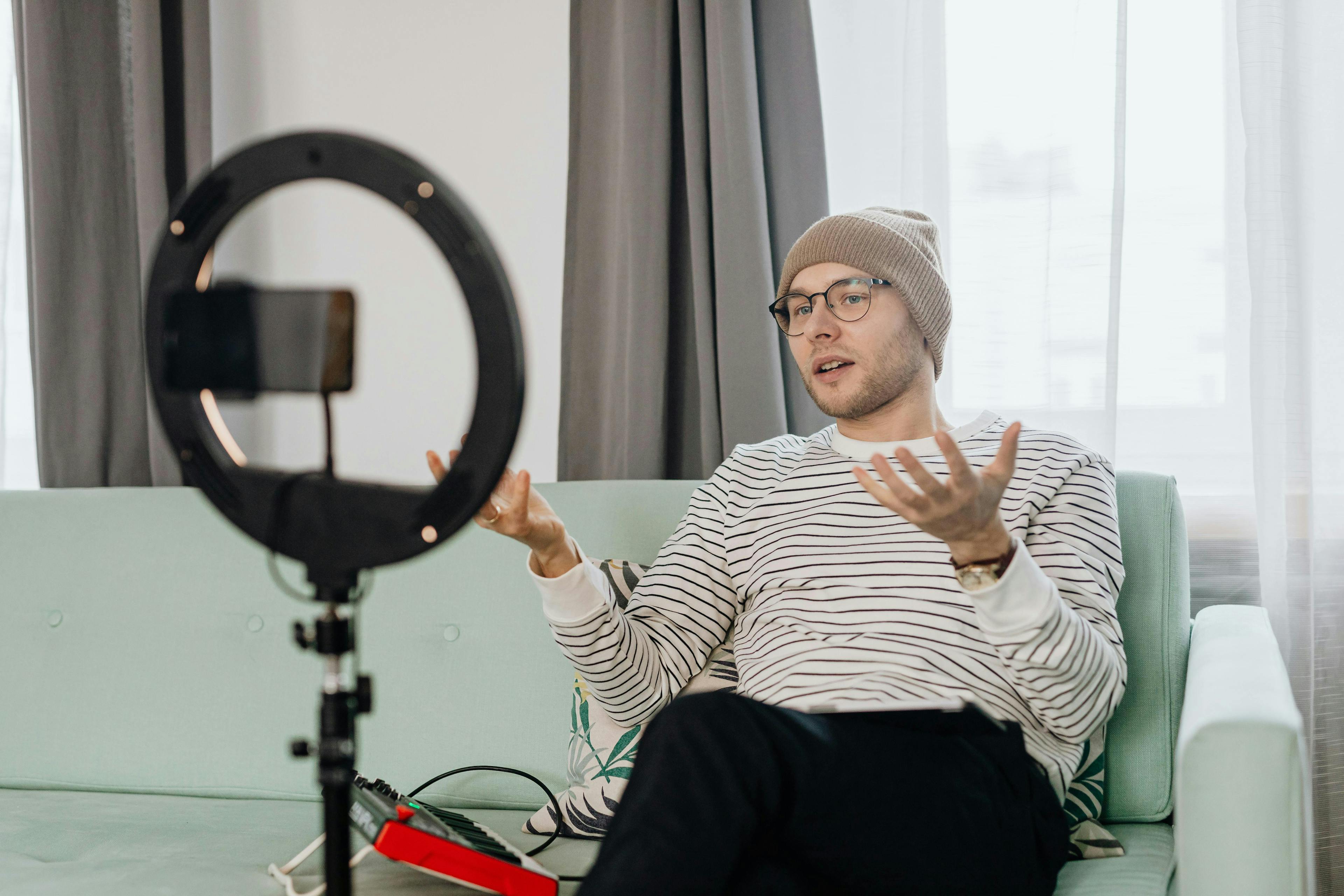 A young man wearing a beanie and glasses is sitting on a light green sofa, speaking animatedly into a smartphone mounted on a tripod with a ring light. The scene suggests he is recording a video, possibly related to Audi marketing. The background features modern curtains and a bright, well-lit room, creating a professional yet casual atmosphere.