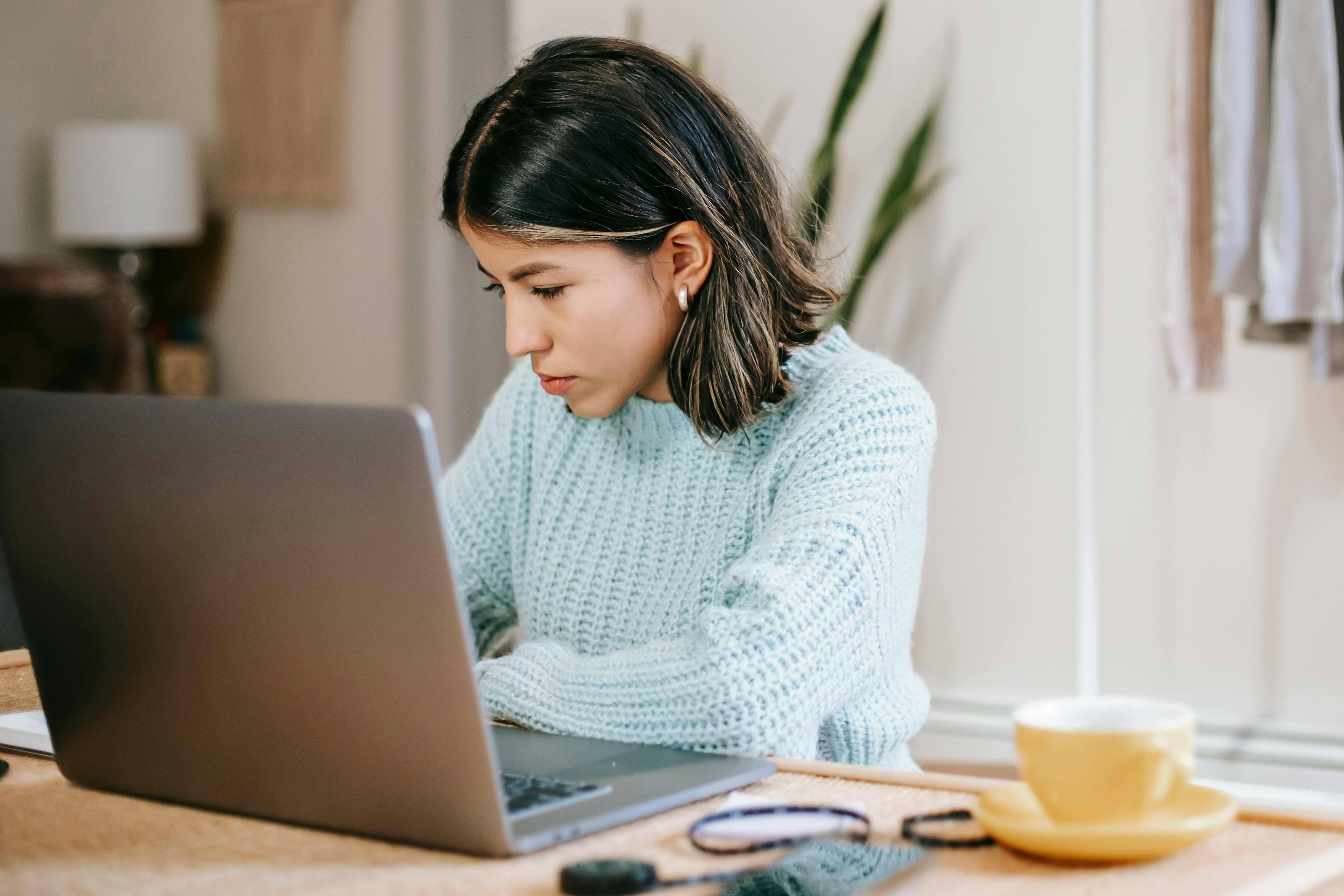 A young woman in a blue sweater, deeply focused on her laptop with a cup of coffee nearby, analyzing data related to market events this week.