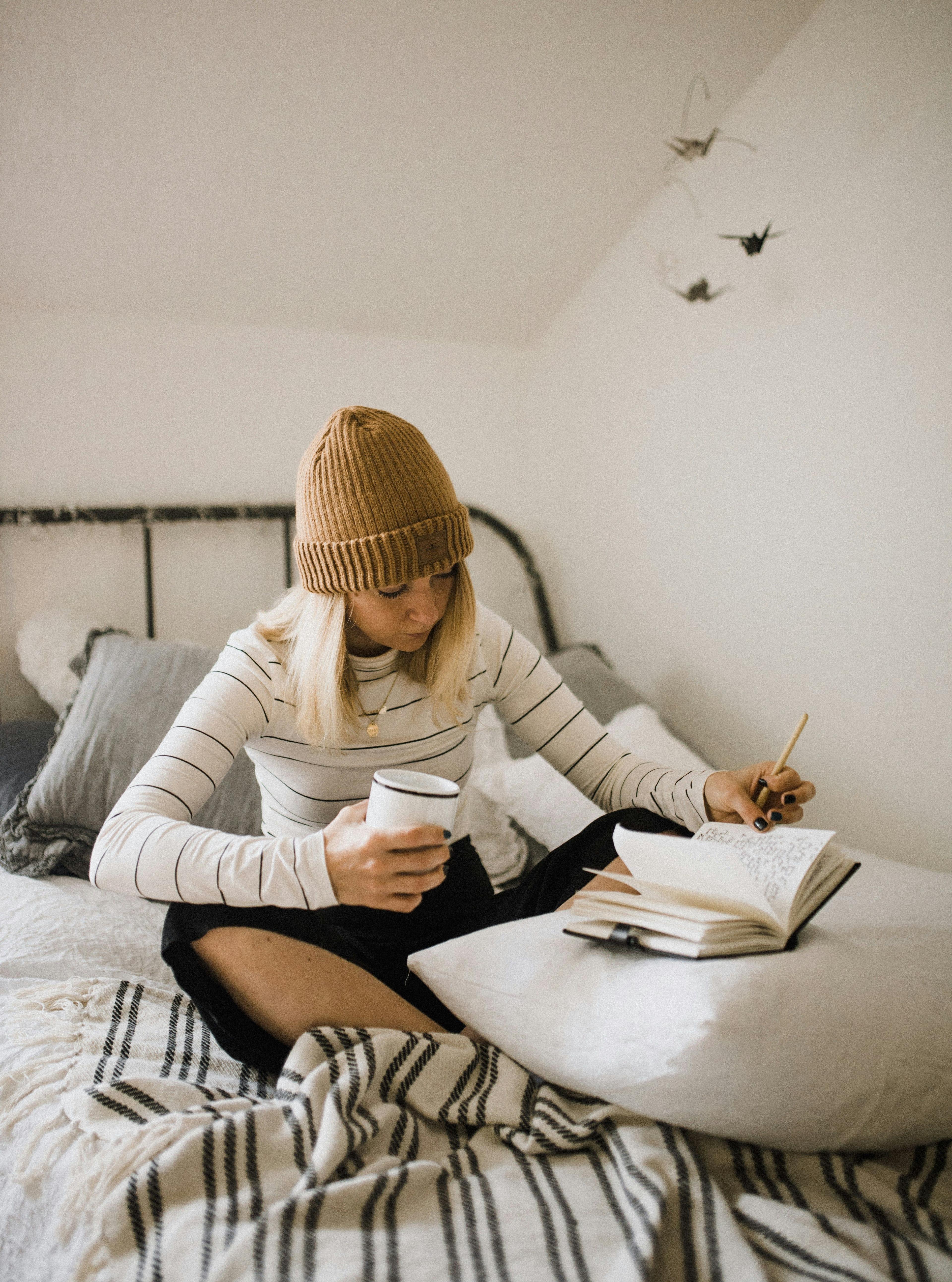 A woman sitting on a bed, wearing a beanie, writing in a notebook while holding a cup, suggesting a relaxed and creative workspace.