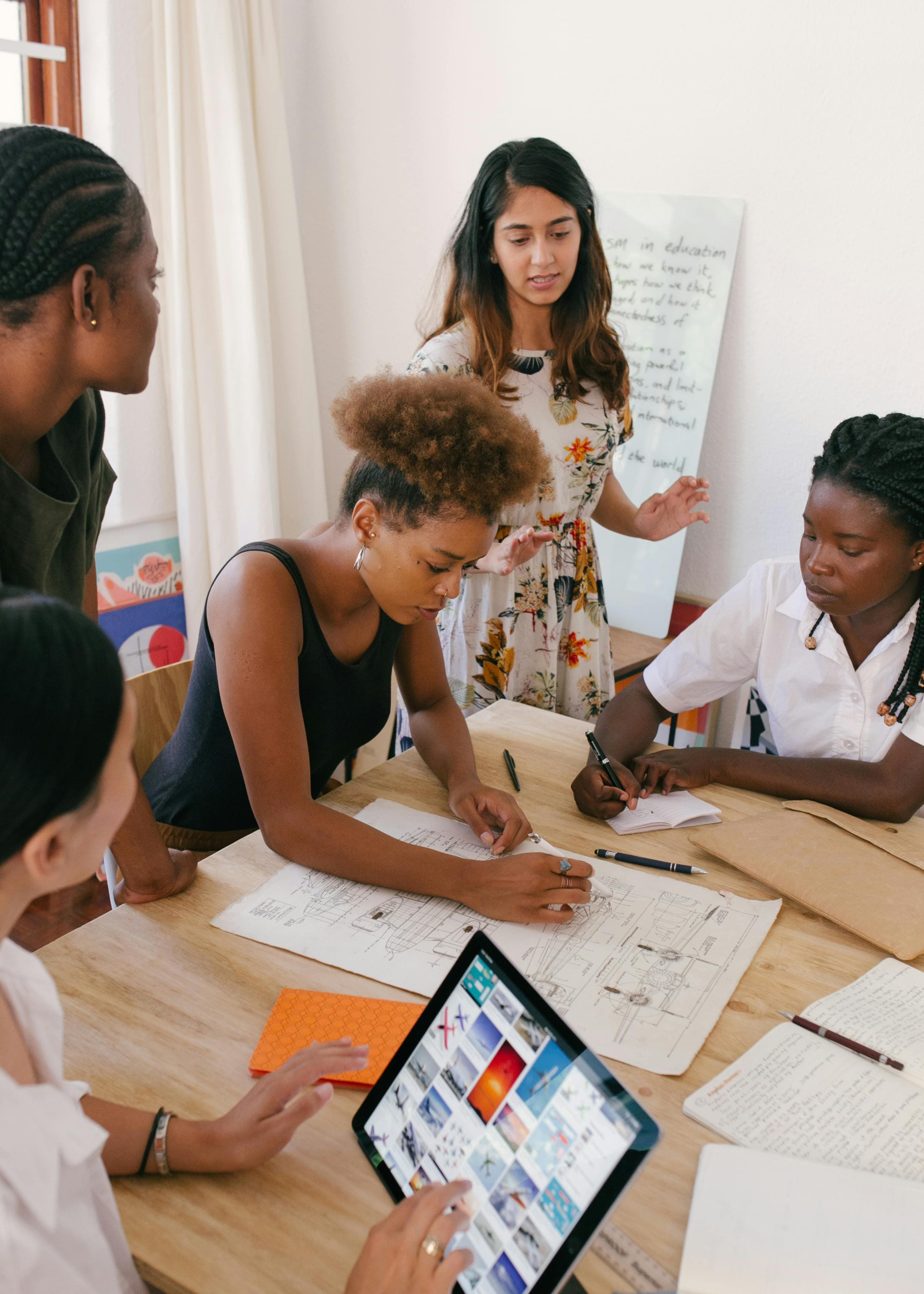 A diverse team of young professionals gathered around a table, working on sketches and designs with one person using a tablet to browse images. This represents a collaborative approach to creating optimized web pages using an AI landing page generator for effective digital solutions.