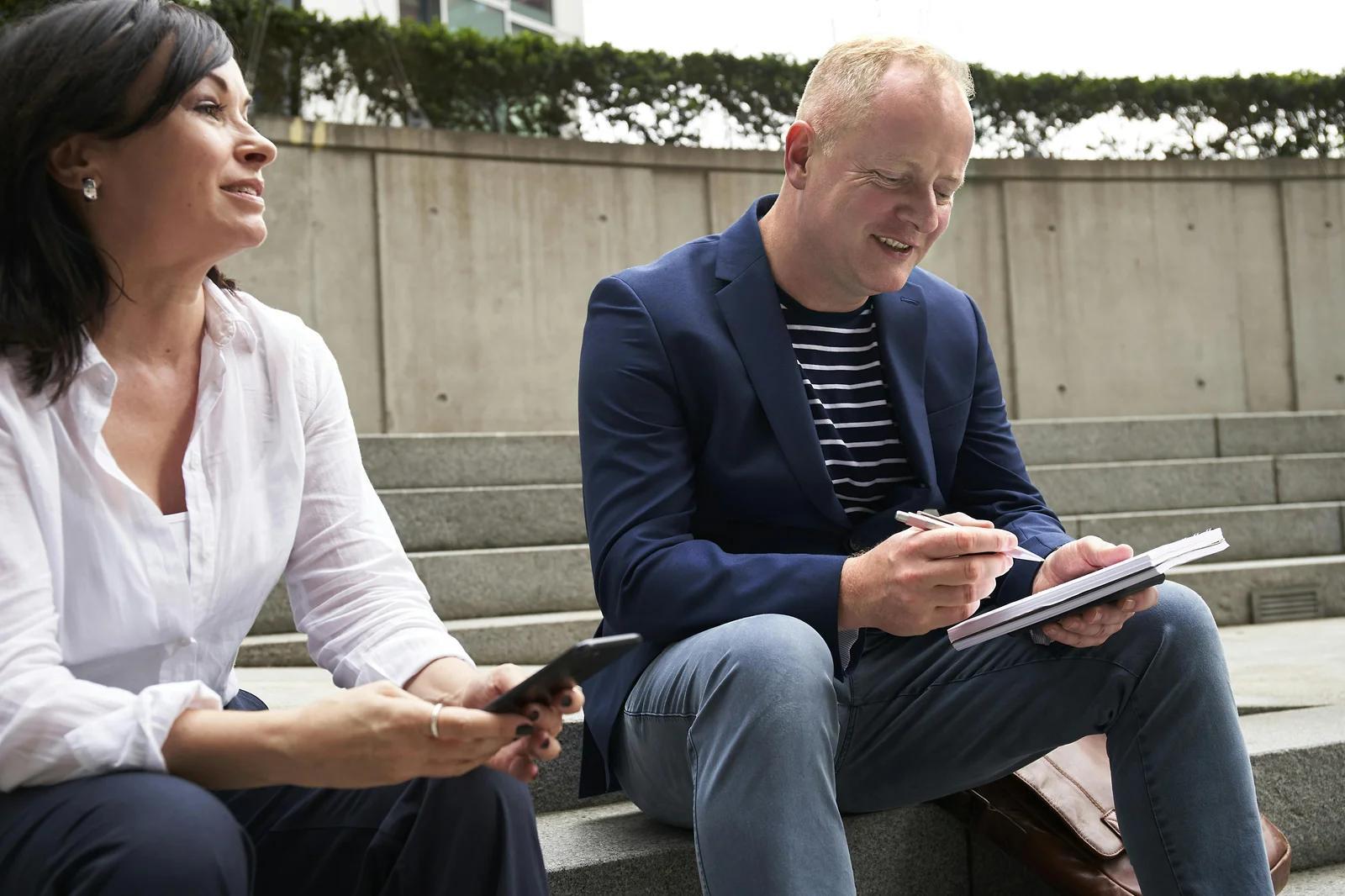 Two people sitting on outdoor steps, discussing and taking notes. One person is holding a phone, and the other is writing in a notebook, possibly brainstorming a video marketing strategy in a collaborative setting.