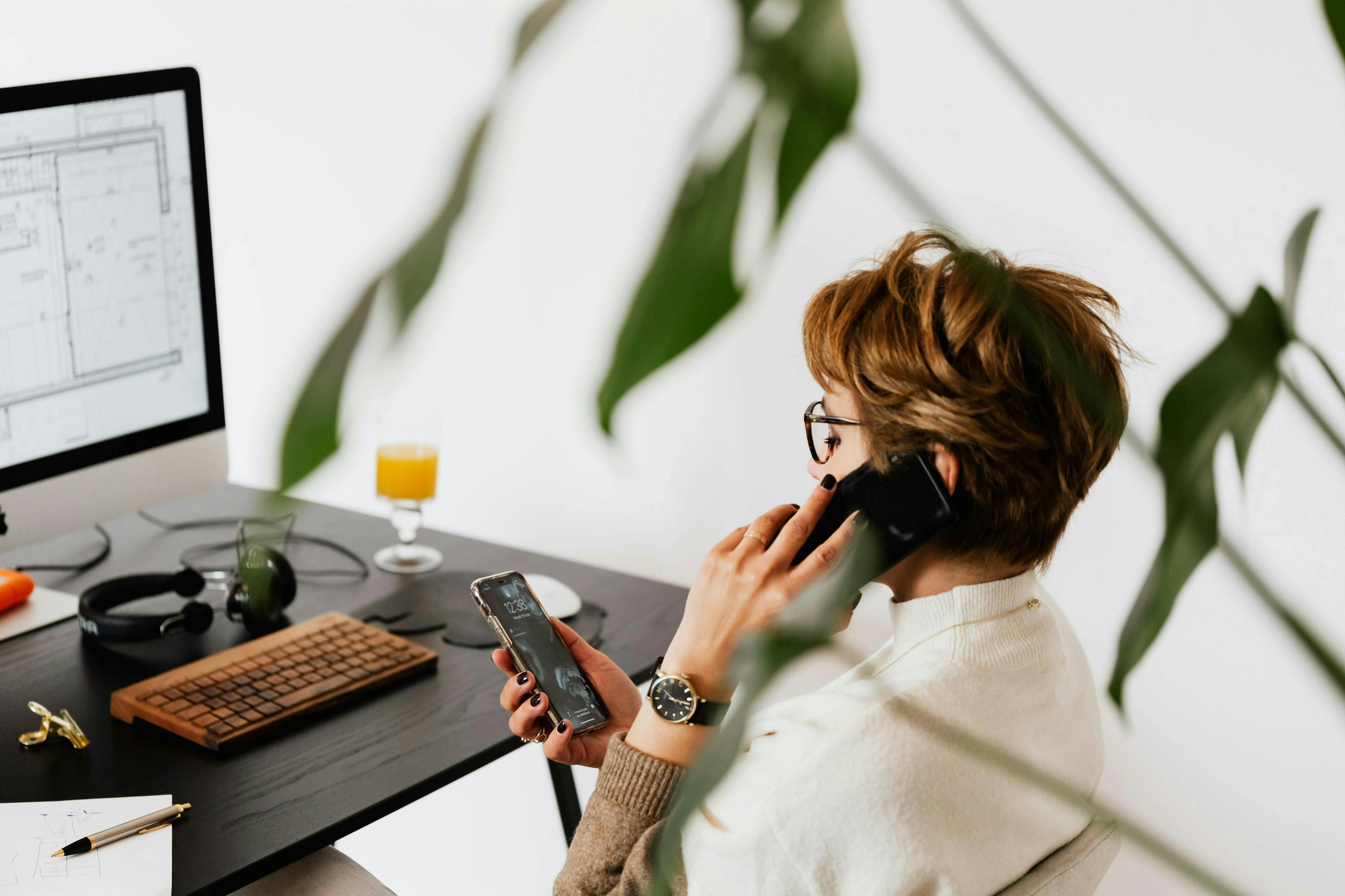 A woman multitasking at her desk, speaking on the phone while checking her smartphone, with a computer screen displaying architectural plans in the background. How to market your coaching business.