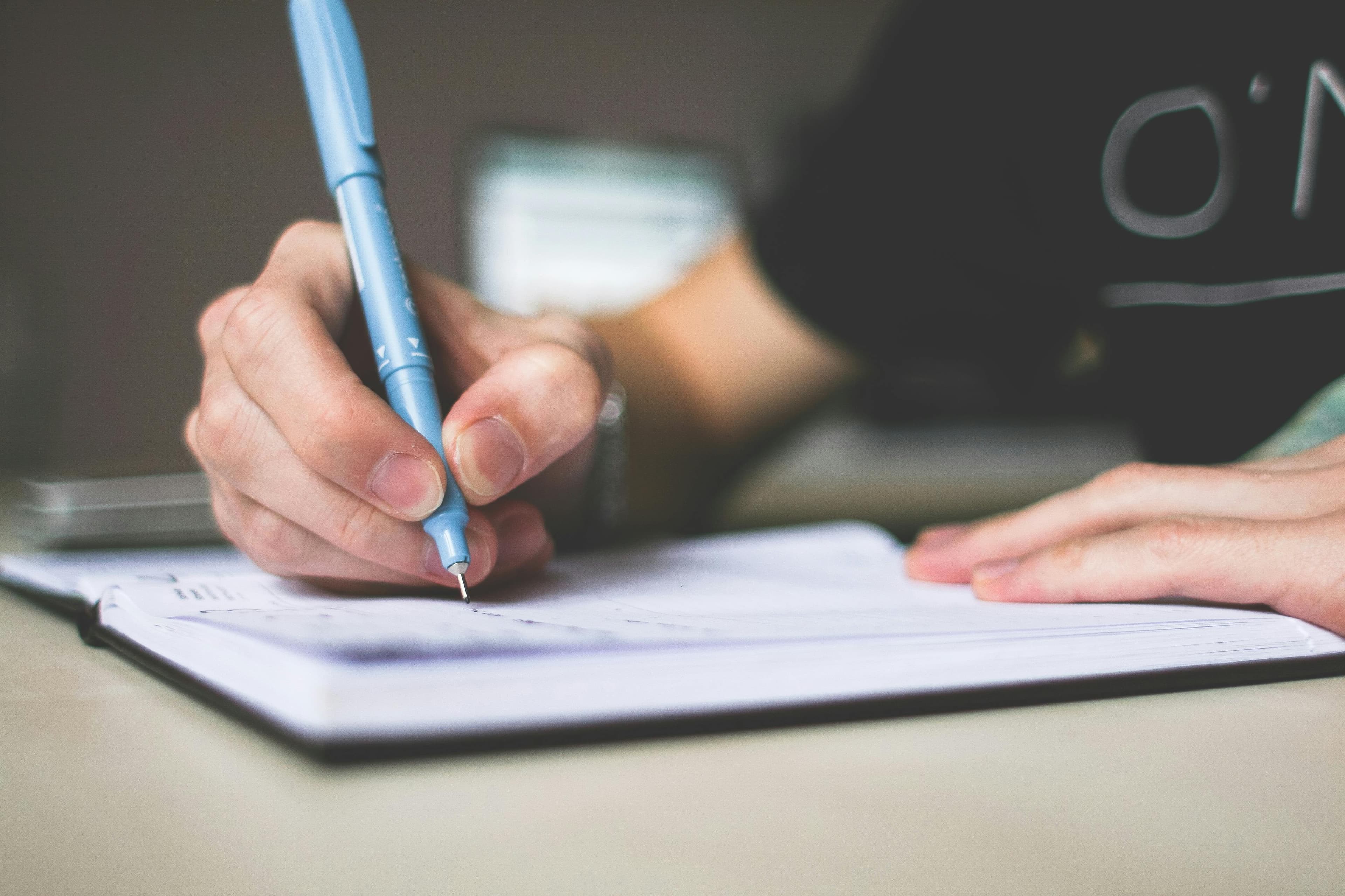 A close-up shot of a hand holding a blue pen, writing in a notebook, signifying documenting the insights and data collected from SEO A/B testing.