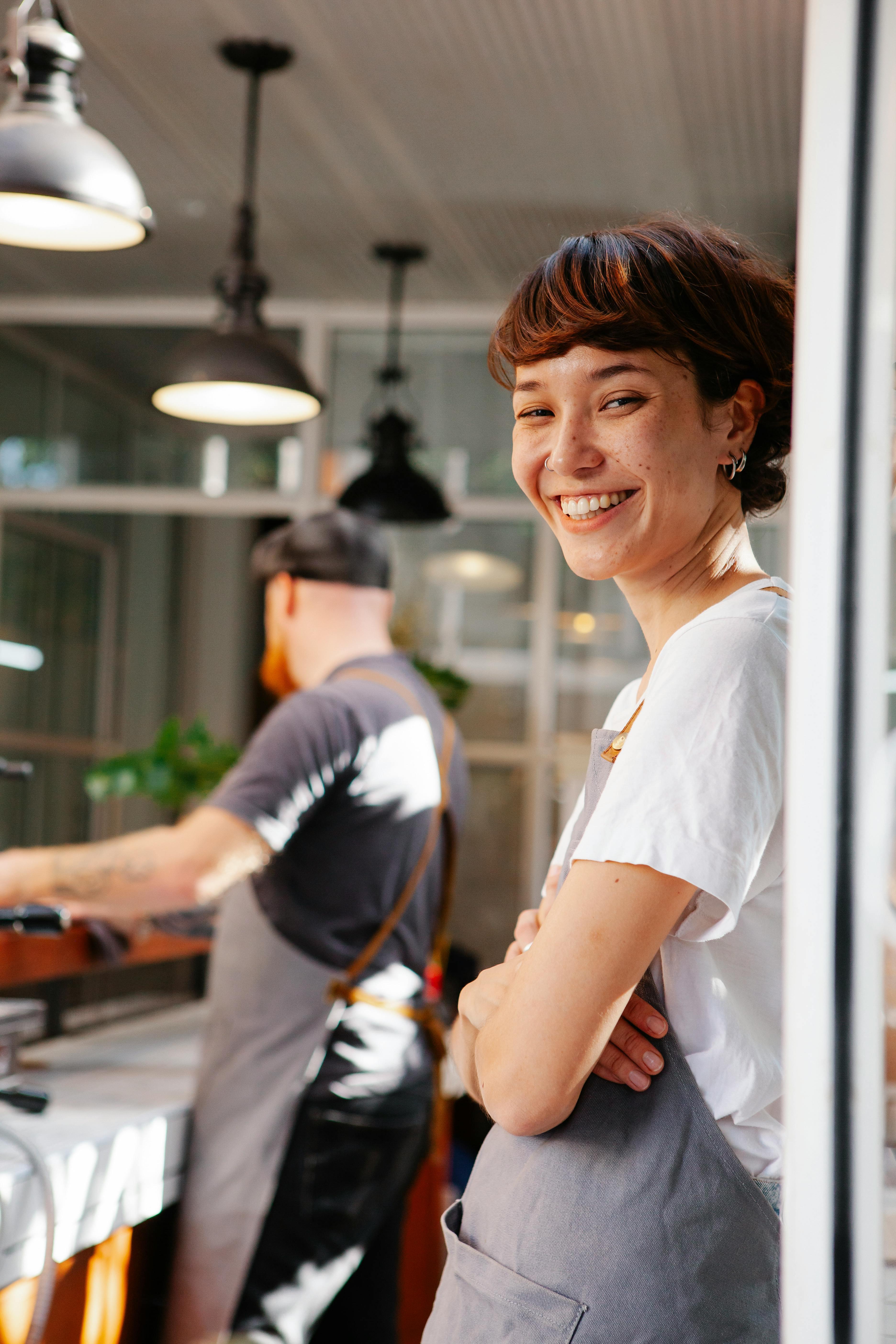 A smiling barista wearing an apron in a cafe environment, showcasing a positive and friendly atmosphere, relevant to UX jobs that focus on creating user-friendly and approachable interfaces.