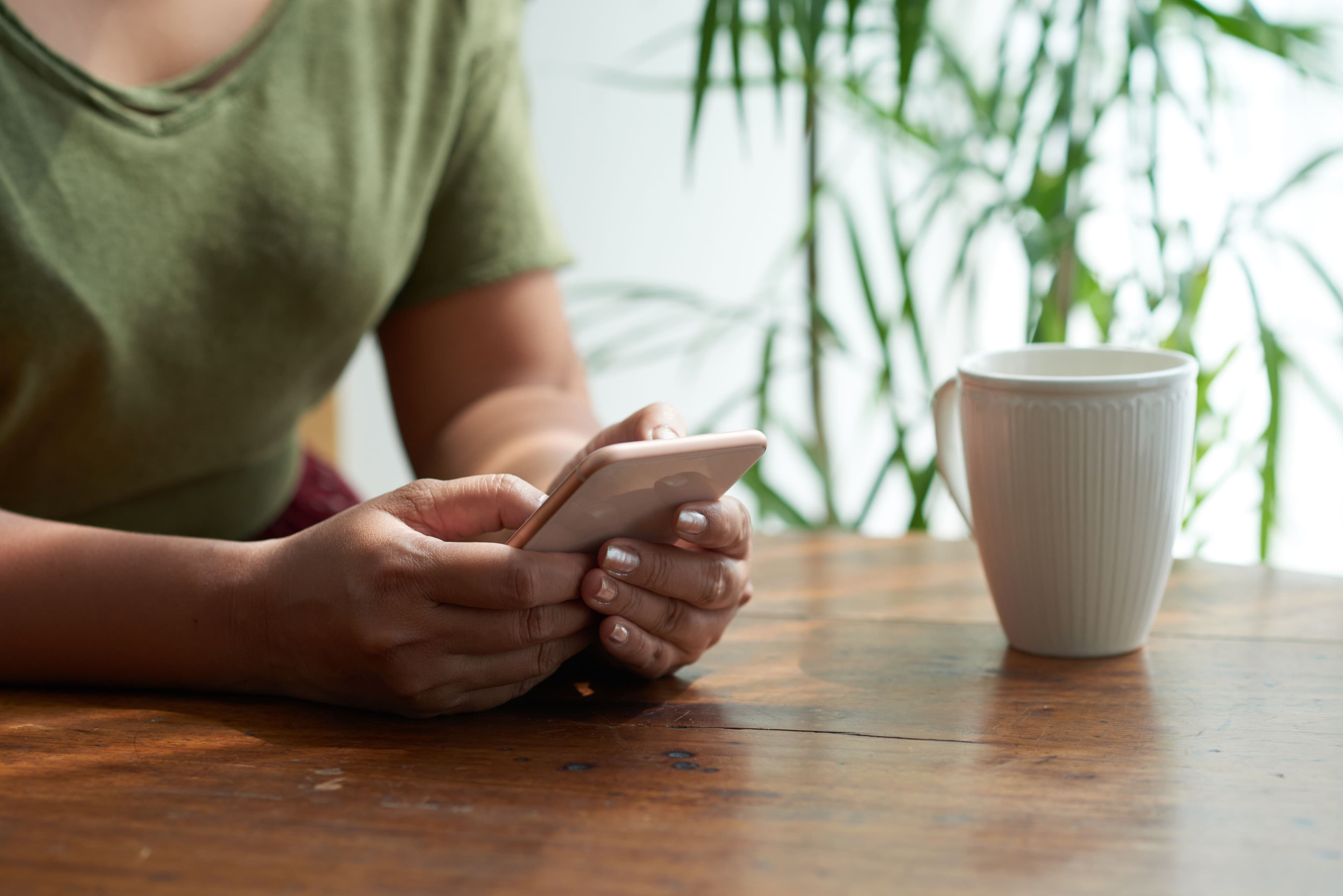 A person sitting at a wooden table in a cozy cafe, using a smartphone, possibly researching small business marketing consulting strategies, with a white coffee mug nearby.