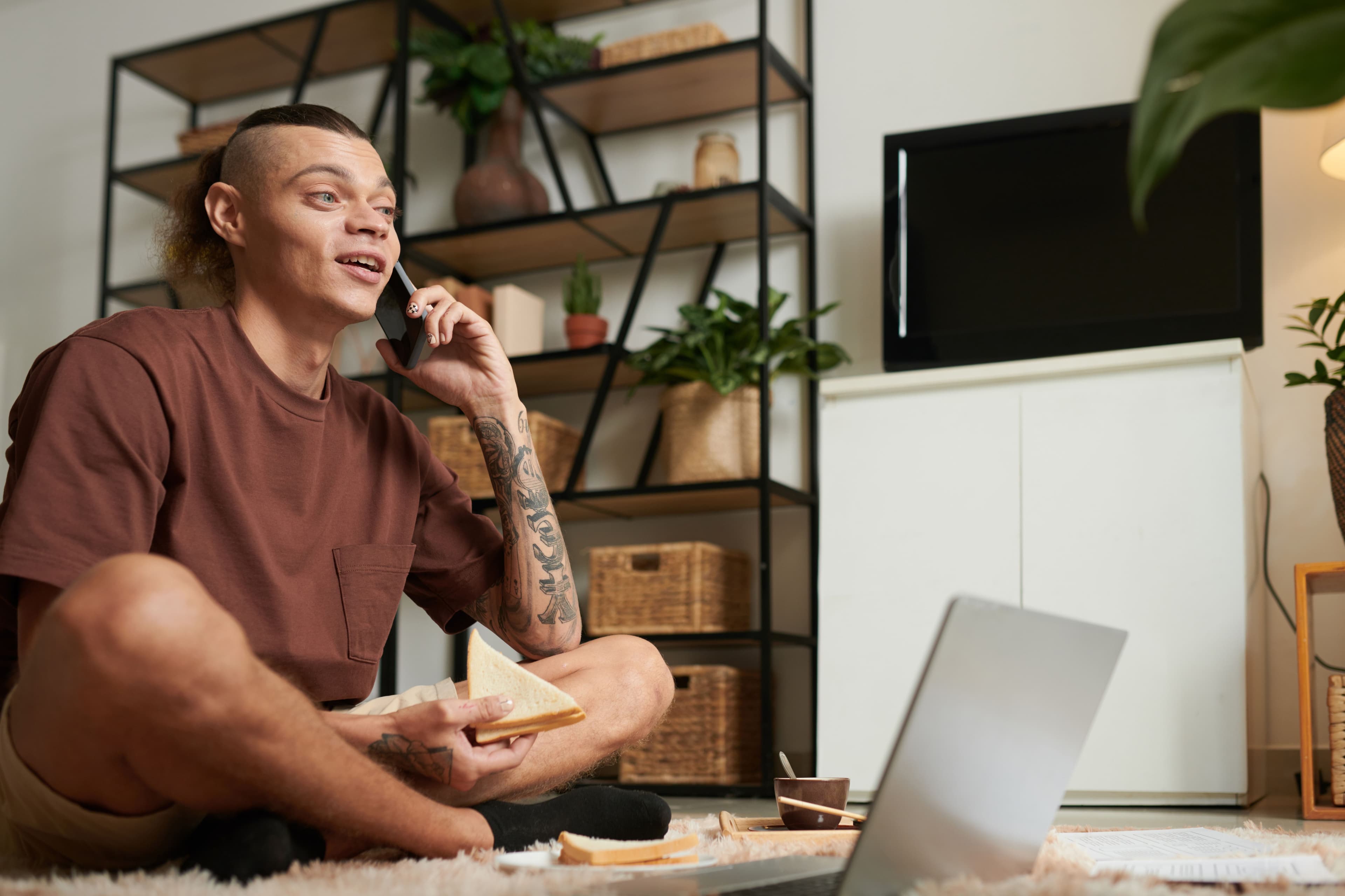 A man with tattoos sitting on the floor, holding a phone and a sandwich, with a laptop open nearby, symbolizing casual work-from-home setups while considering social media ad cost.