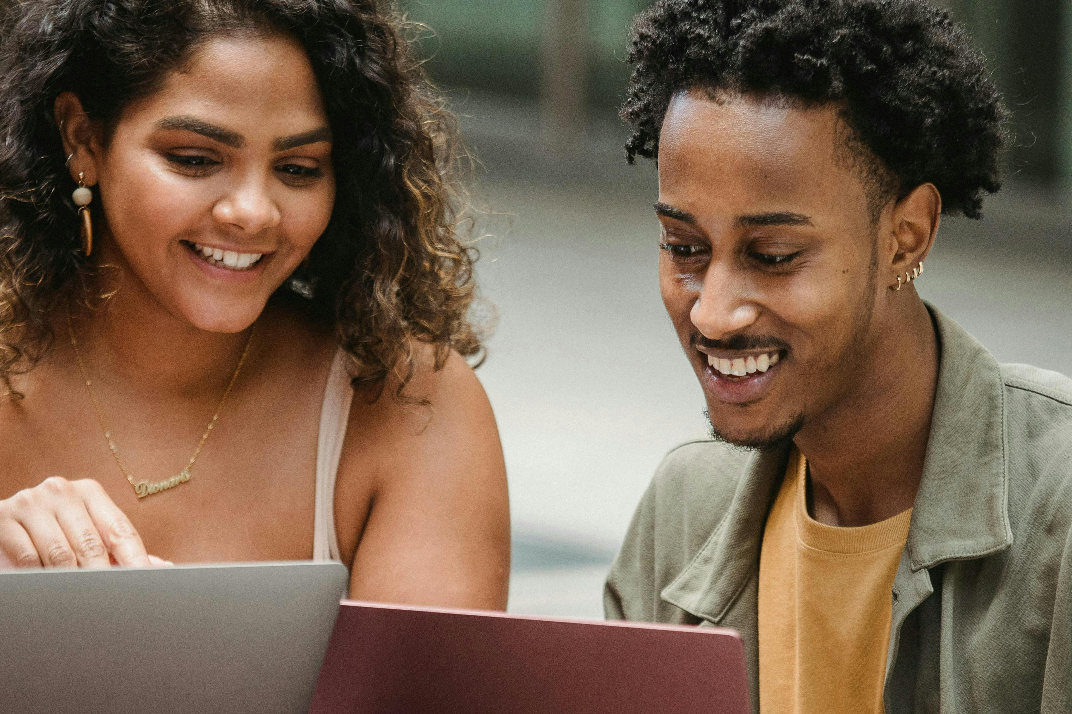 Two individuals happily collaborating while looking at laptops. The woman on the left, wearing a beige top and gold hoop earrings, is smiling and engaged in the conversation. The man on the right, dressed in a green jacket and yellow shirt, is also smiling as they work together. They appear to be discussing strategies or content related to email marketing, creating a positive and productive atmosphere for their project.