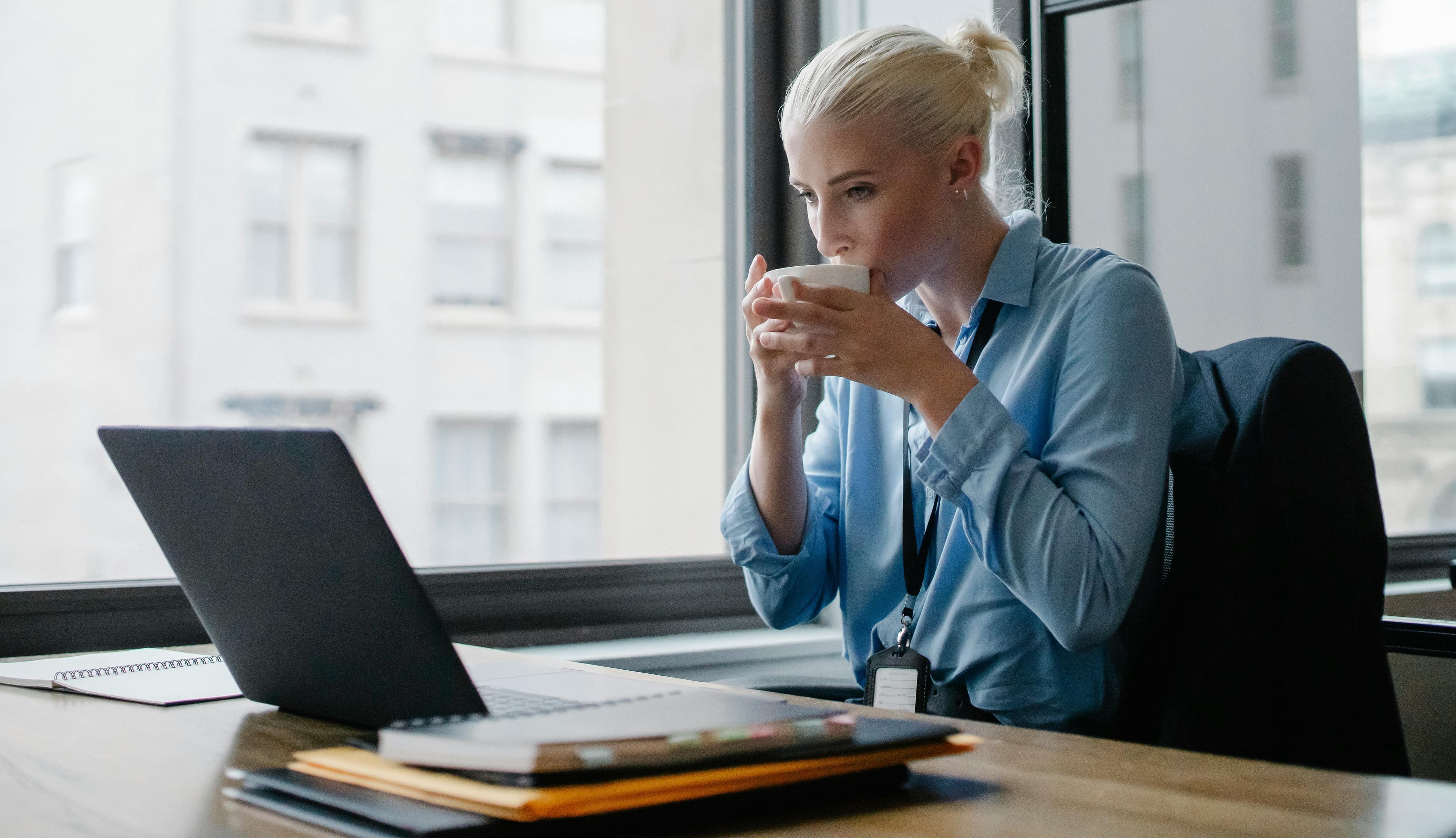 A woman with blonde hair tied in a bun, dressed in a light blue blouse, is sitting at a desk by a large window, drinking from a white cup. She is working on a laptop with notebooks and documents spread out on the desk, suggesting she is engaged in tasks related to email marketing jobs. The office environment is bright and professional, with a view of city buildings through the window, indicating a productive workday.