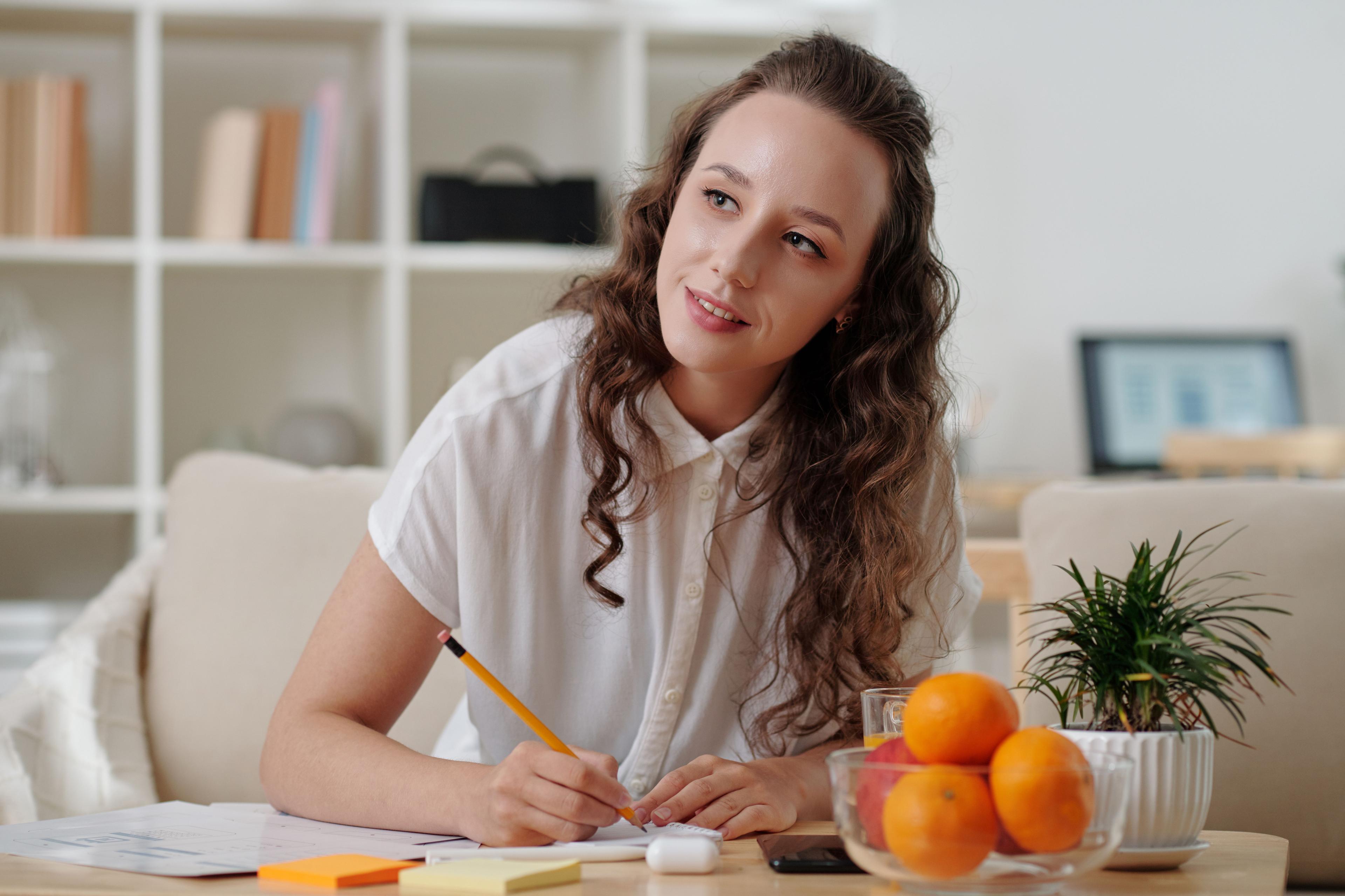  A woman with curly hair, wearing a white shirt, is sitting at a table, writing notes on paper with colorful sticky notes and a bowl of oranges nearby. She appears thoughtful and focused in a well-organized, calm indoor setting, symbolizing another dataset in the "sklearn train test split."