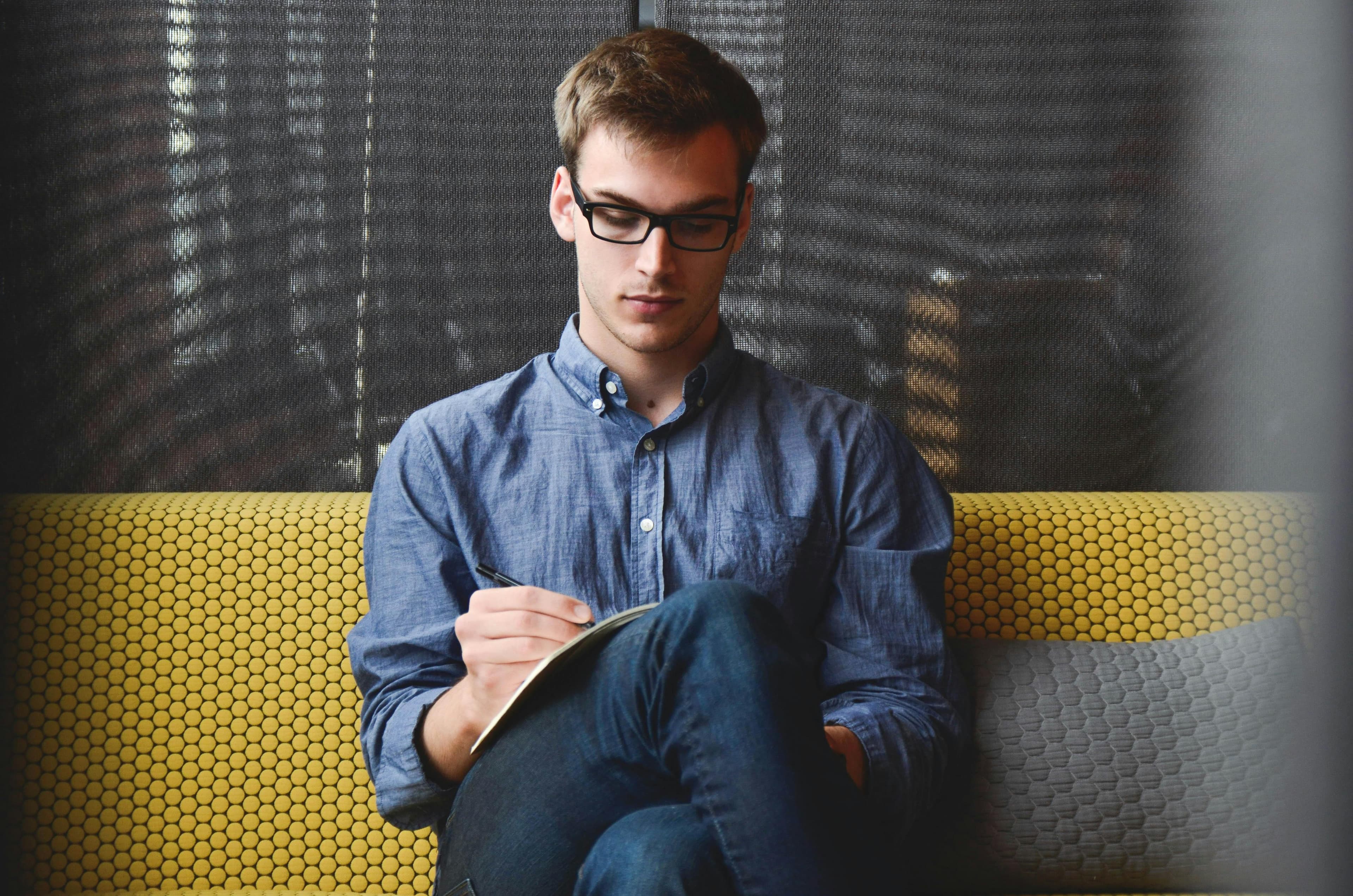 A person in a blue shirt and glasses sits on a couch writing in a notebook, concentrating on his notes, symbolizing the planning phase of SEO A/B testing.