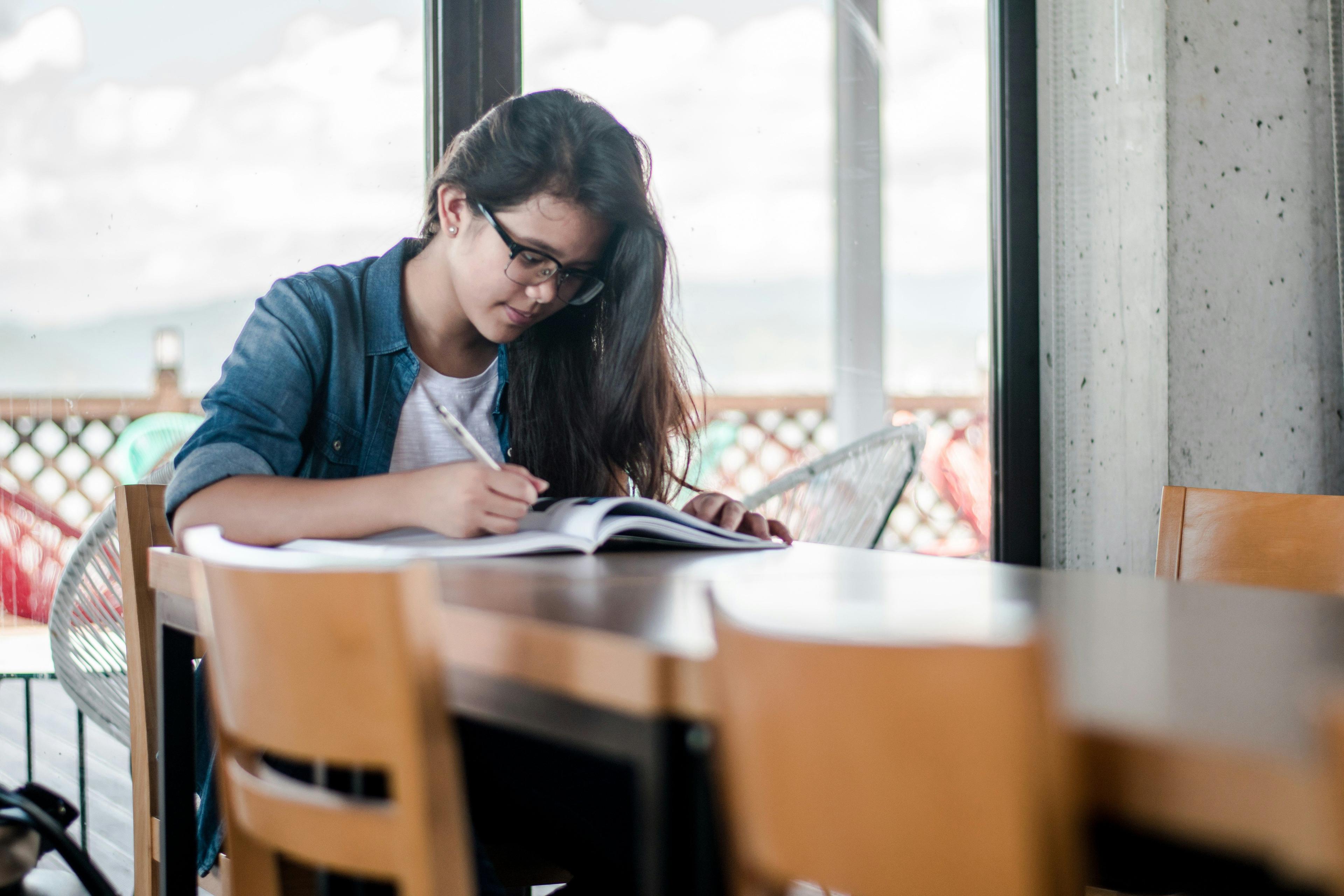 A student studying at a desk by a window, immersed in her notes and research, possibly planning video marketing content for an academic project.