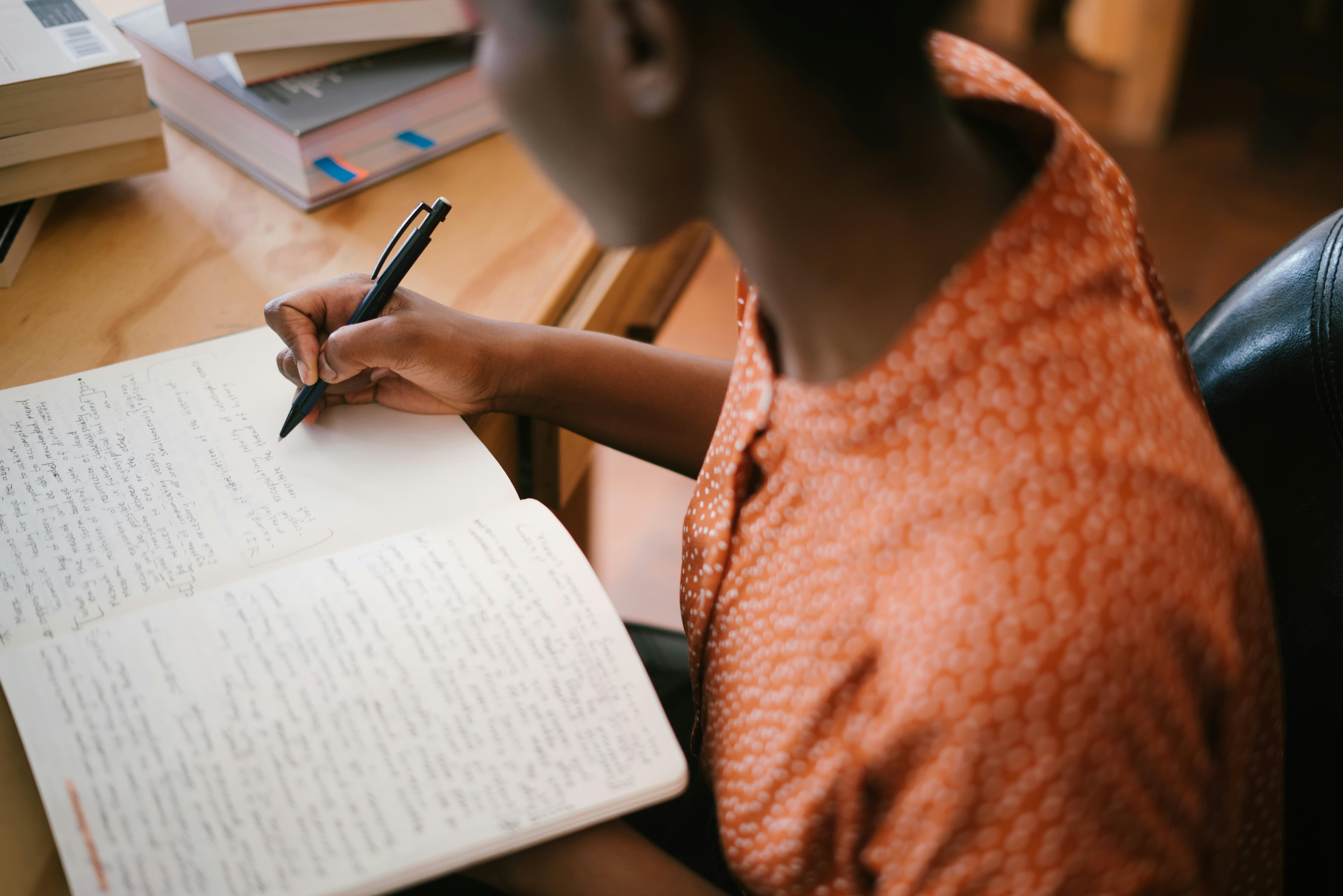 A close-up of a person in an orange shirt writing notes in a notebook at a desk, symbolizing the importance of planning and organizing ideas before using an AI landing page generator to streamline the web design process.