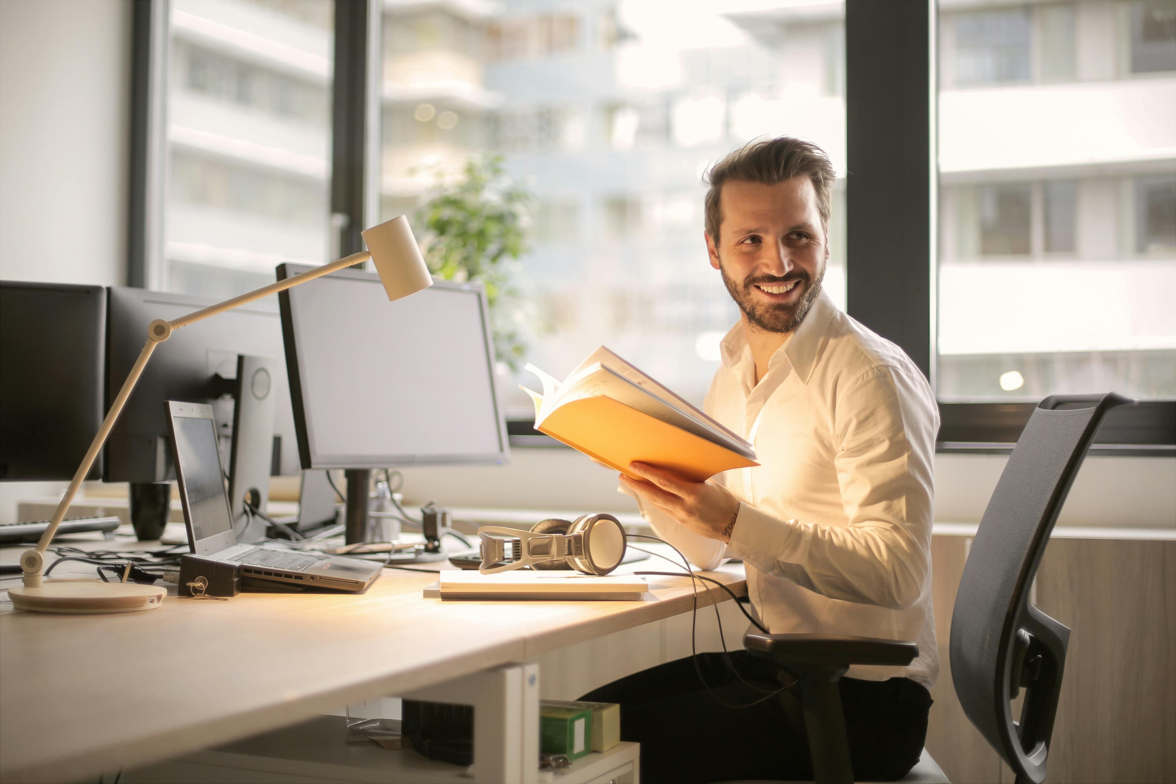 A smiling man in an office setting holds an open book, facing a desk with multiple monitors and equipment. This image conveys a welcoming and productive environment, suitable for a "lead form template" targeted at professional consultations or educational resources.