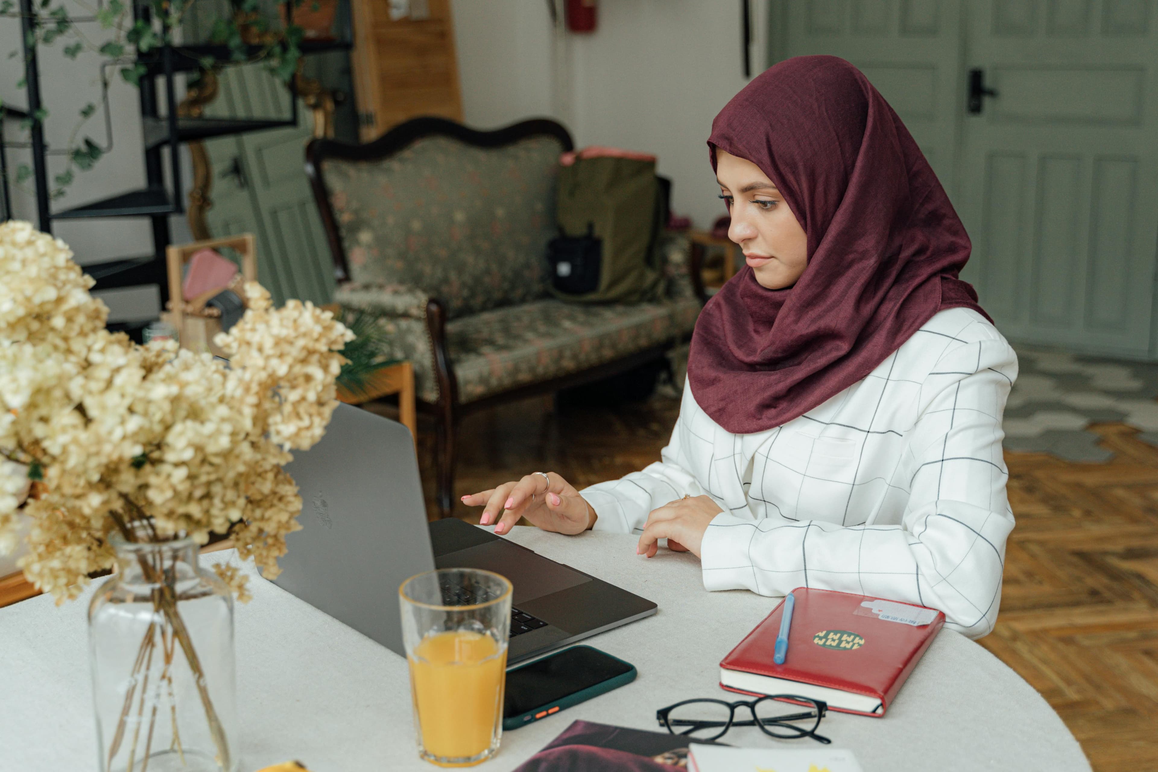 A woman in a maroon hijab working on a laptop in a cozy home setting, with a notebook, glasses, and orange juice on the table. This reflects a focus on remote work and strategic planning for conversion optimization in digital marketing.