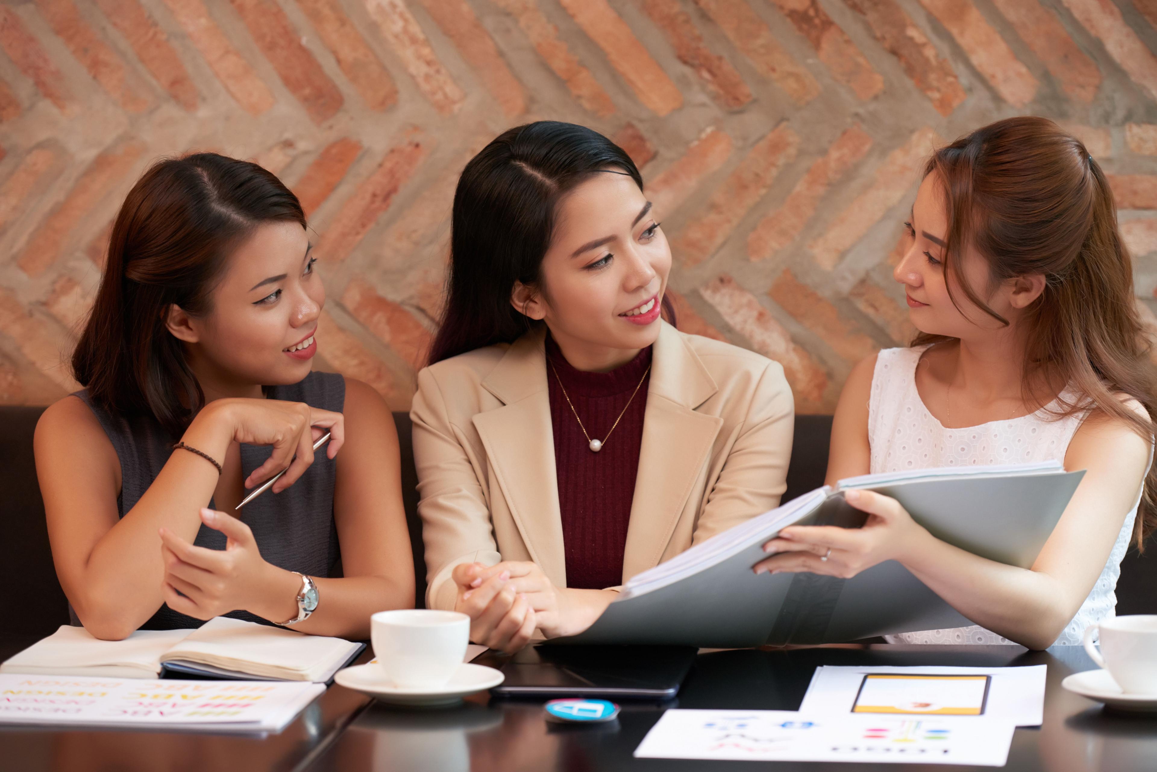 Three businesswomen engaged in a lively discussion, reviewing documents over coffee in a casual cafe setting. Alt Text: "Group of professional women strategizing social media ad campaigns.