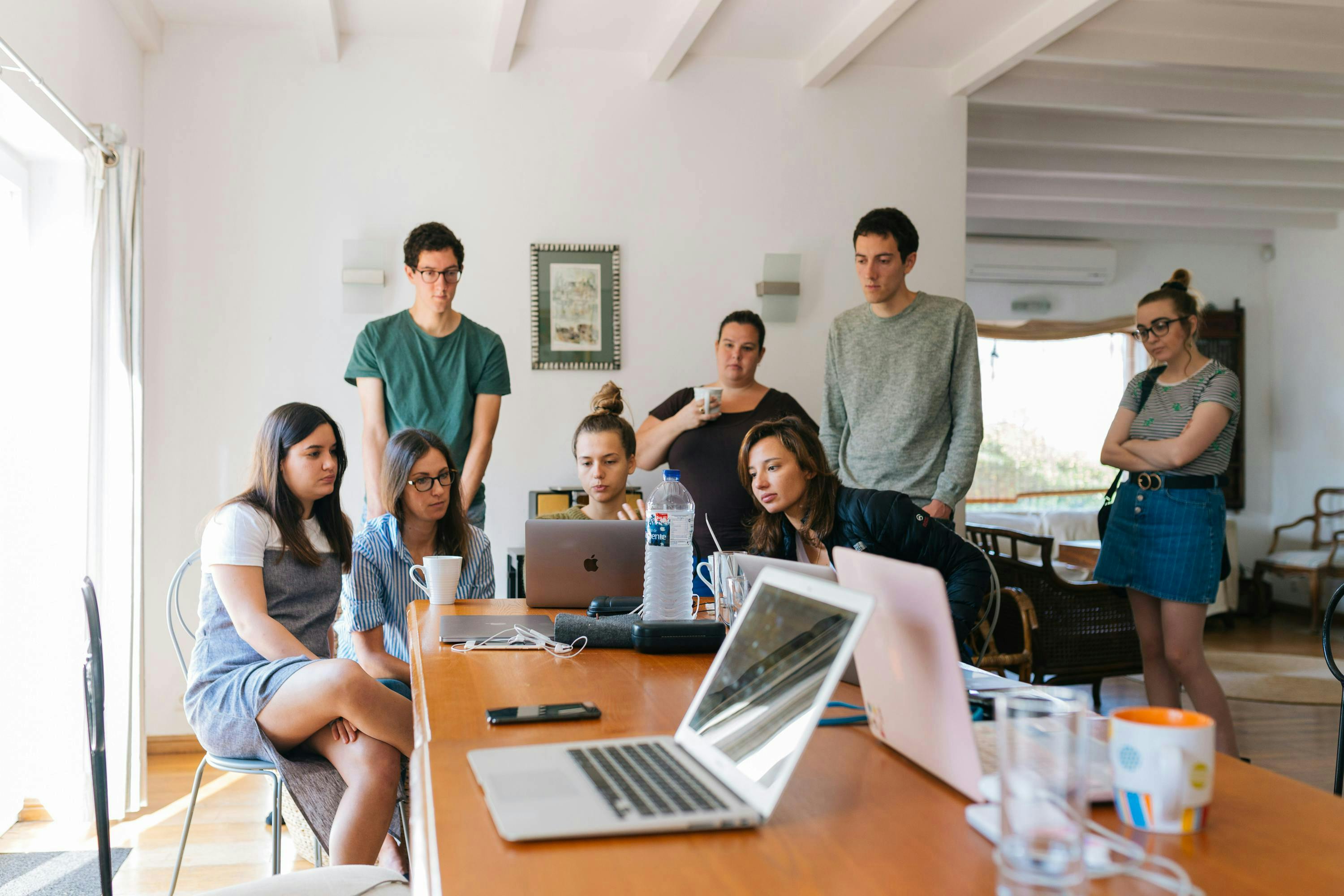 A diverse group of individuals gathered around a table, focusing on a laptop screen during a collaborative meeting. This image is ideal for depicting User Experience (UX) Researcher Jobs, highlighting teamwork and collective brainstorming.