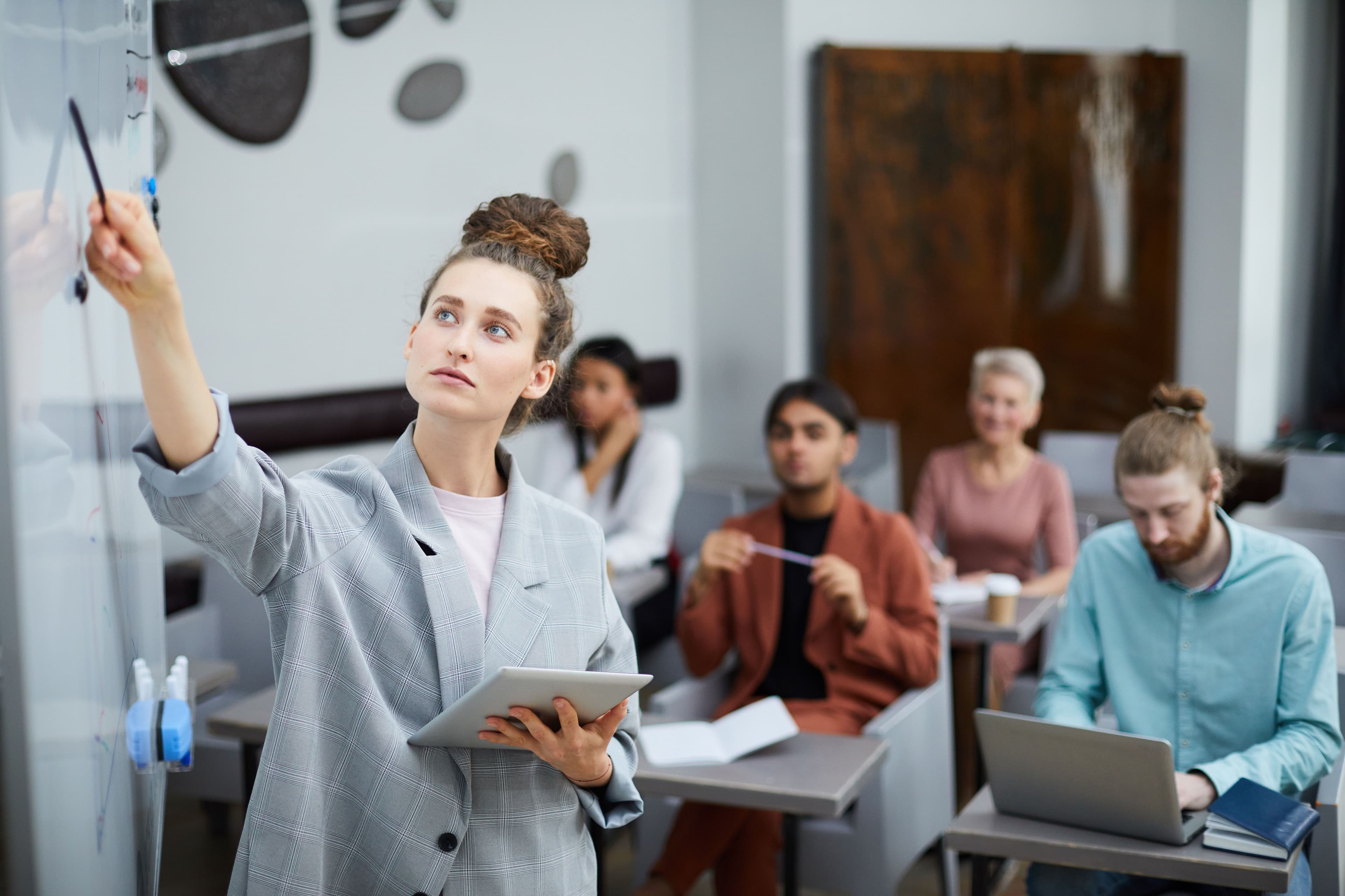 A young businesswoman pointing at a whiteboard while holding a tablet, surrounded by attentive colleagues in a modern conference room, indicating a team discussion about a "social media marketing strategy."