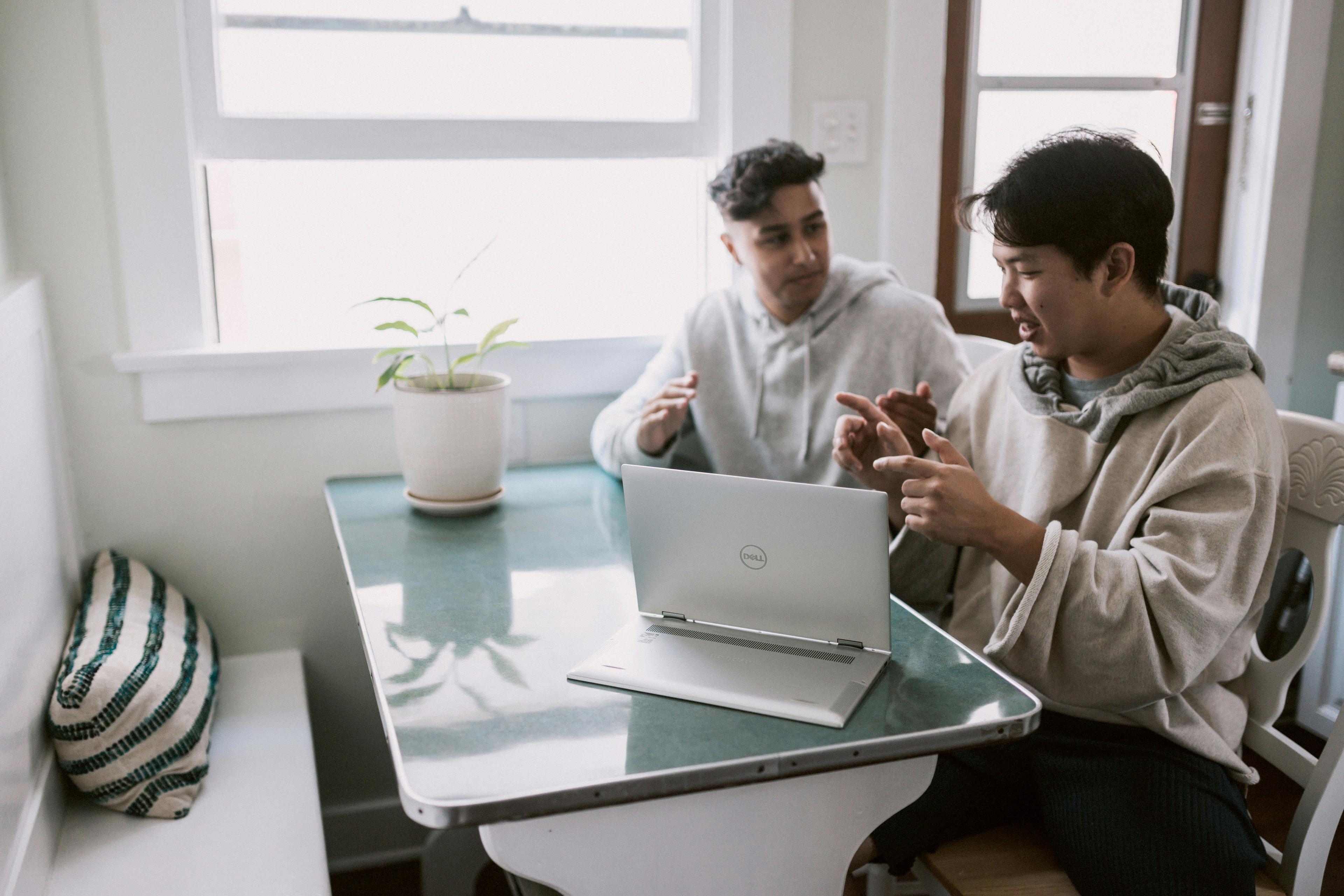 Two people sitting at a small kitchen table, discussing and collaborating over a laptop. The setting suggests a home office or casual workspace, reflecting the collaborative aspect of "small business consulting marketing."