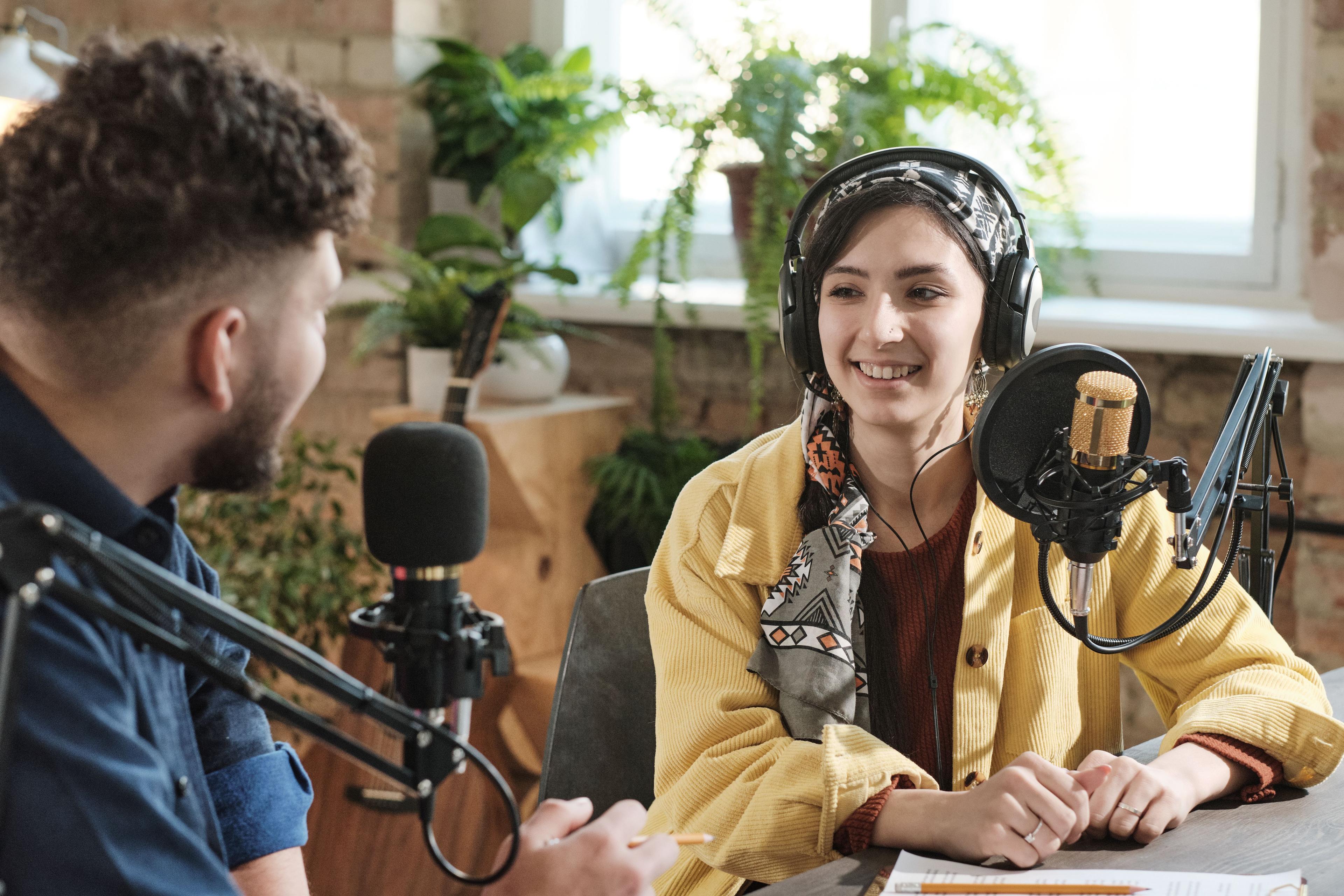 A man and woman in a radio studio setting are engaged in conversation. Both wear headphones and speak into microphones, with plants and cozy decor in the background.