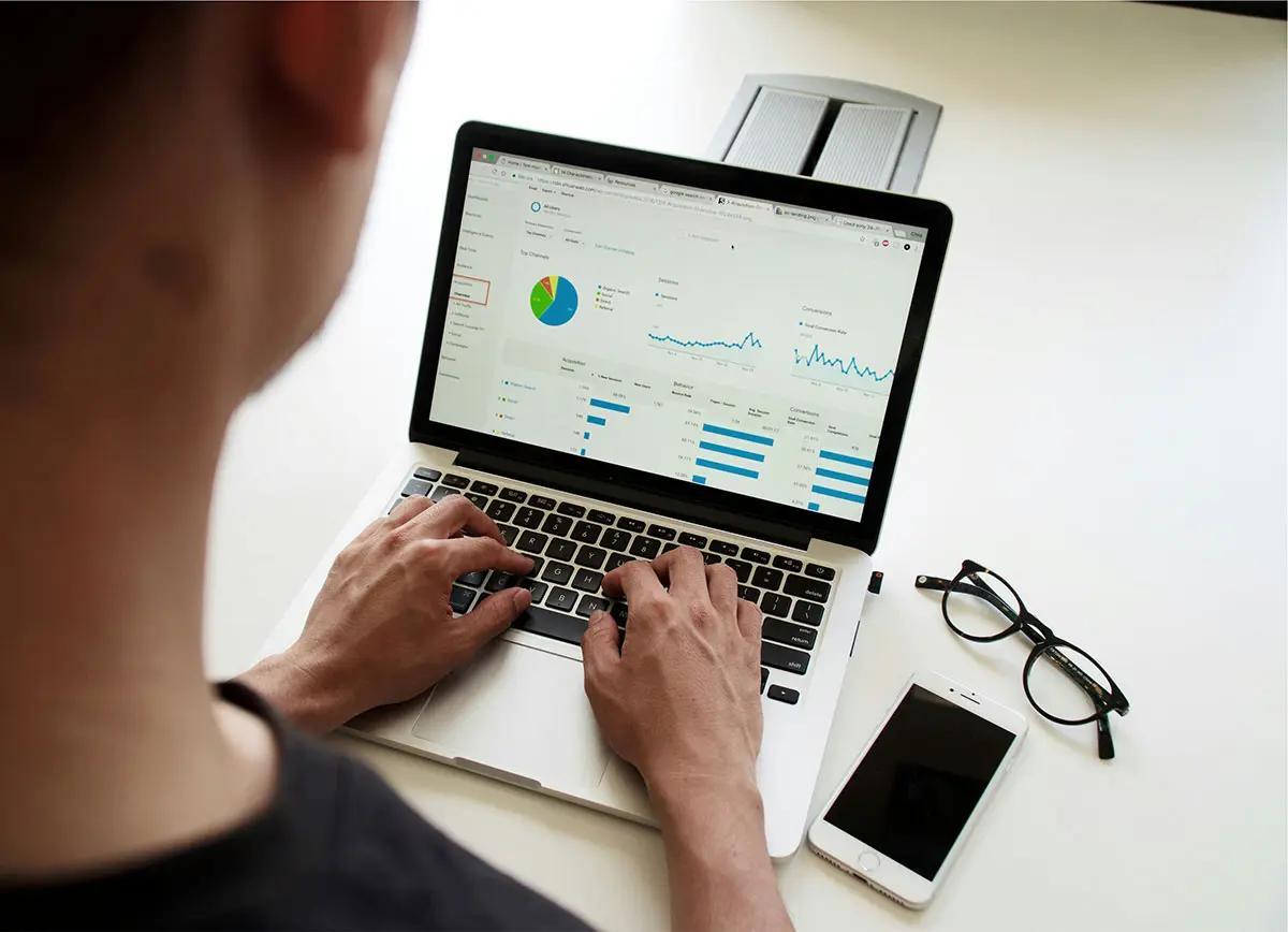 A person is working on a laptop, analyzing data displayed on the screen. The screen shows various graphs and charts, including a pie chart and line graphs, indicating an analysis of performance metrics. Beside the laptop are a pair of glasses and a smartphone, suggesting a focused work environment. This image is suitable for illustrating the concept of "capital markets authority salaries."