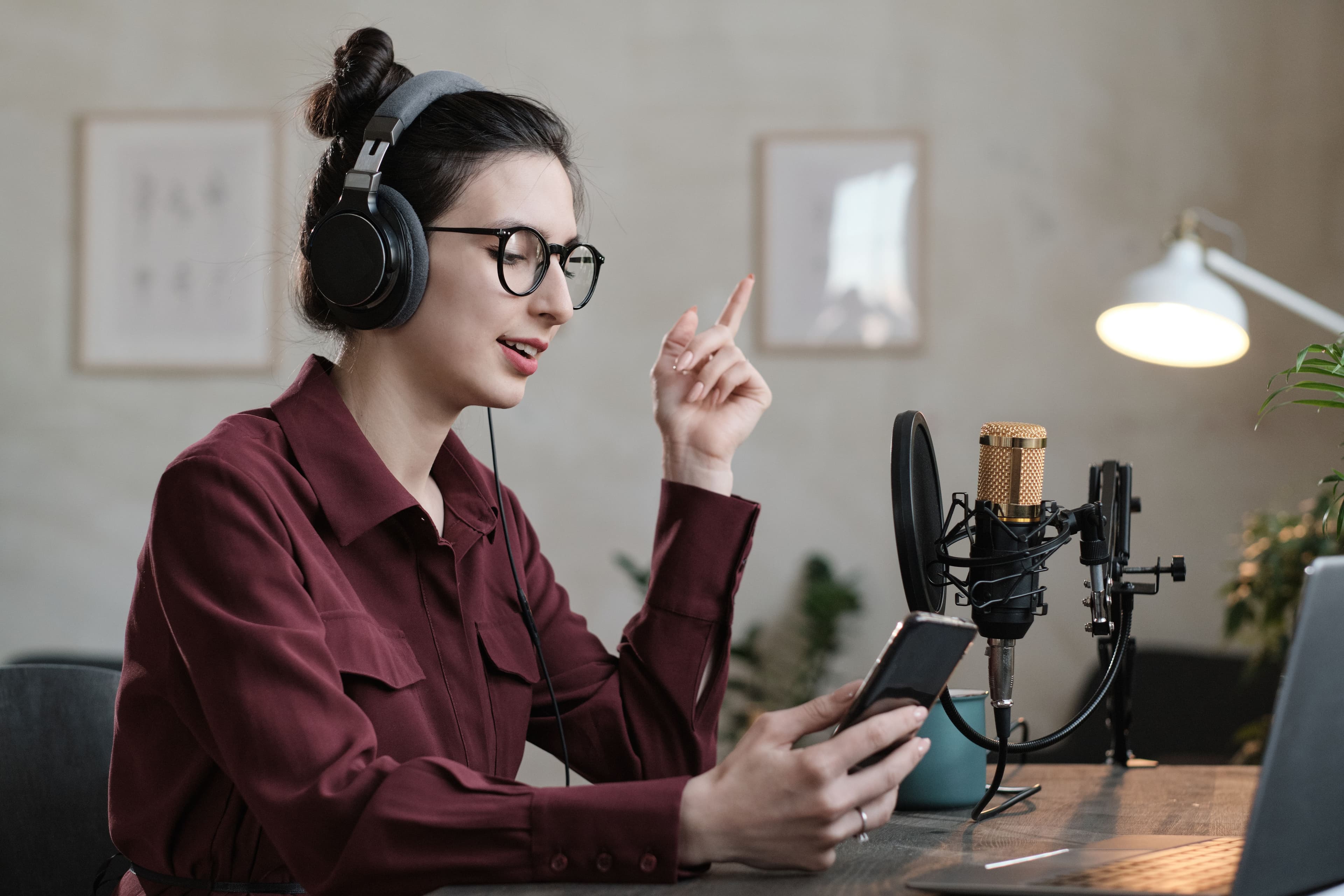 A woman in glasses with headphones on is talking into a microphone while holding a smartphone, illustrating a live radio or podcast recording. A/B testing could be used to compare listener engagement across different hosting styles or content topics. Keywords: a b testing definition, podcast recording, live radio.