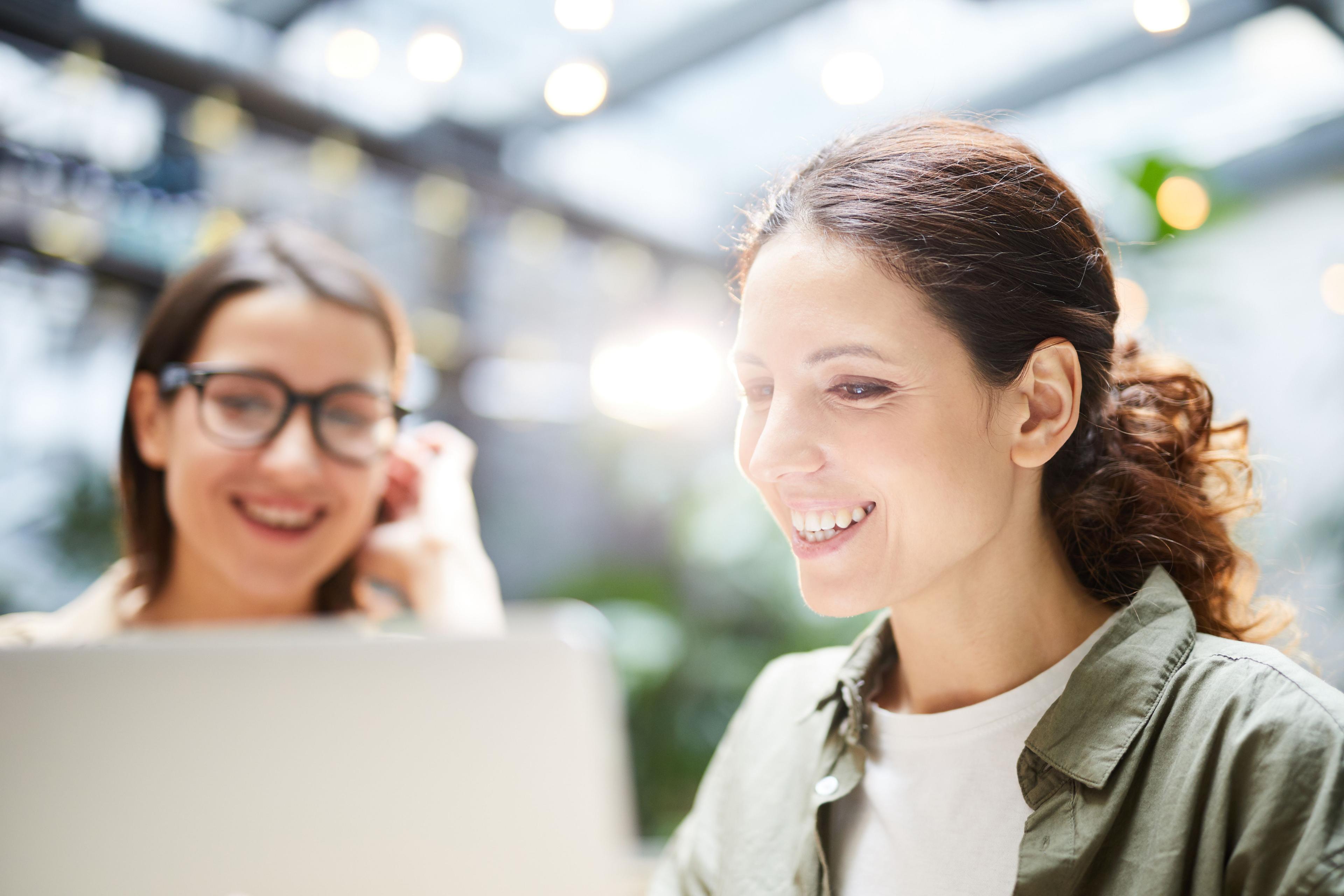 Two women, one with glasses, are smiling while looking at a laptop screen, in a bright, modern workspace. They are collaborating, possibly on research or a digital project.