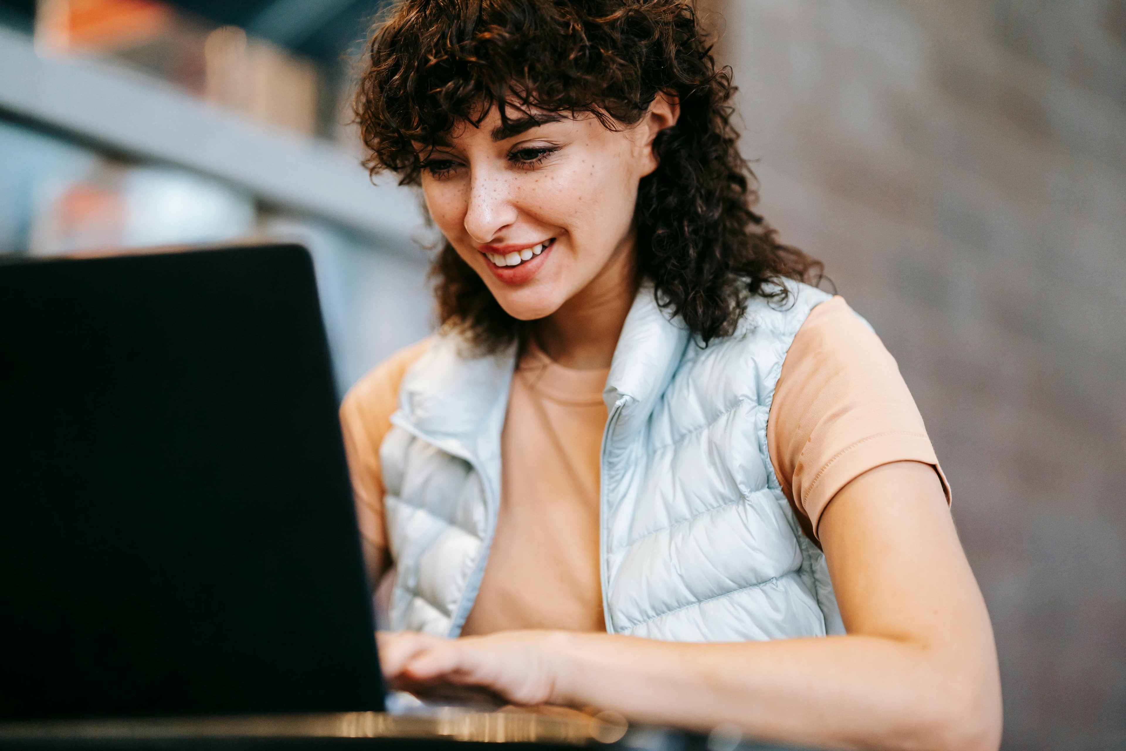 A woman with curly hair and a light vest smiling as she works on her laptop, reflecting the positive outcomes of effective marketing conversion strategies.