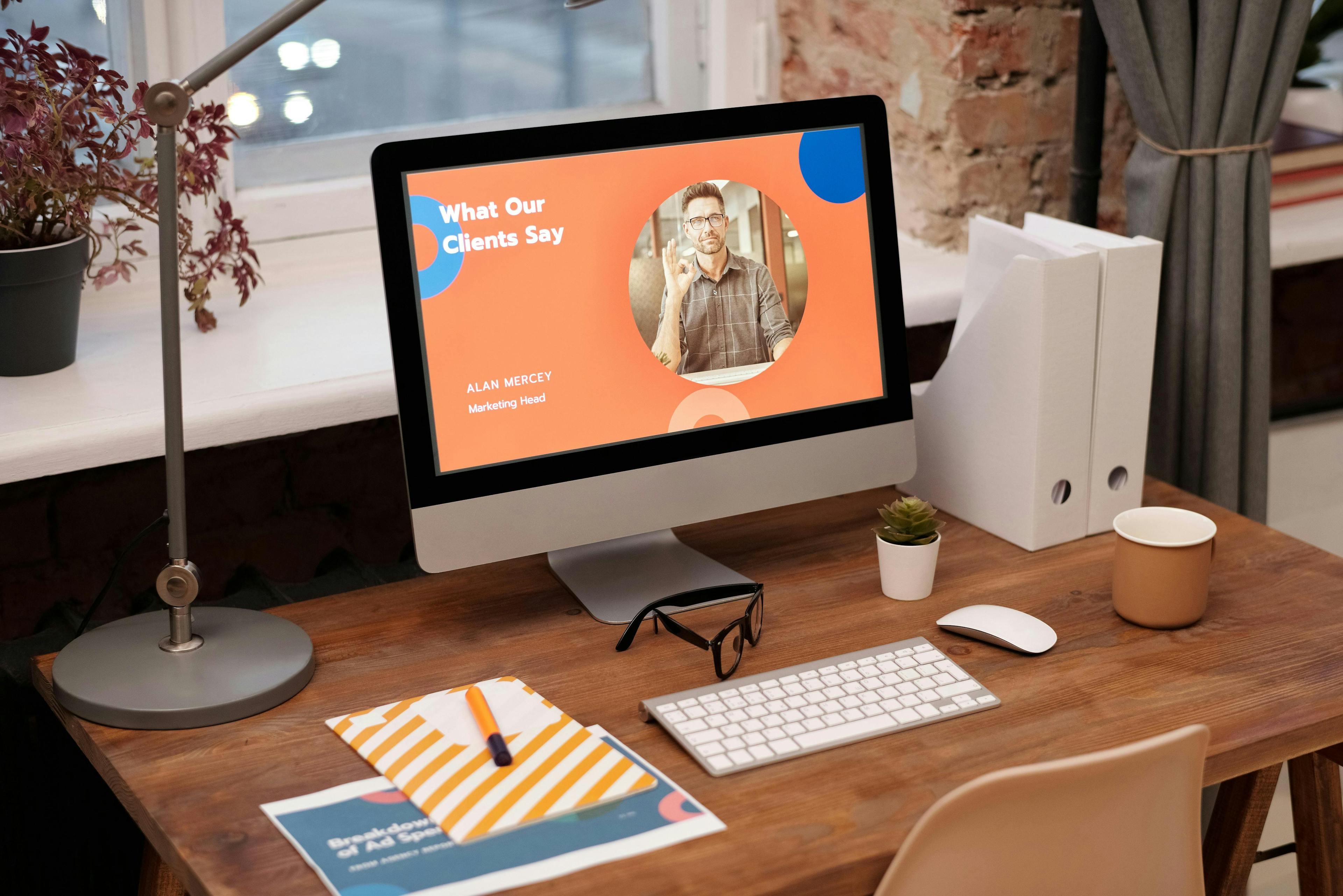 A neatly organized desk featuring a computer displaying a presentation slide titled "What Our Clients Say" with a photo of Alan Mercey, Marketing Head, and a testimonial. The desk also has a lamp, a pair of glasses, a notebook with a pen, a cup, a small potted plant, and white binders. The workspace suggests a professional environment, likely involved in financial services marketing, where client feedback and testimonials are being reviewed or prepared for a presentation. The background includes a window and a brick wall, adding to the cozy office ambiance.