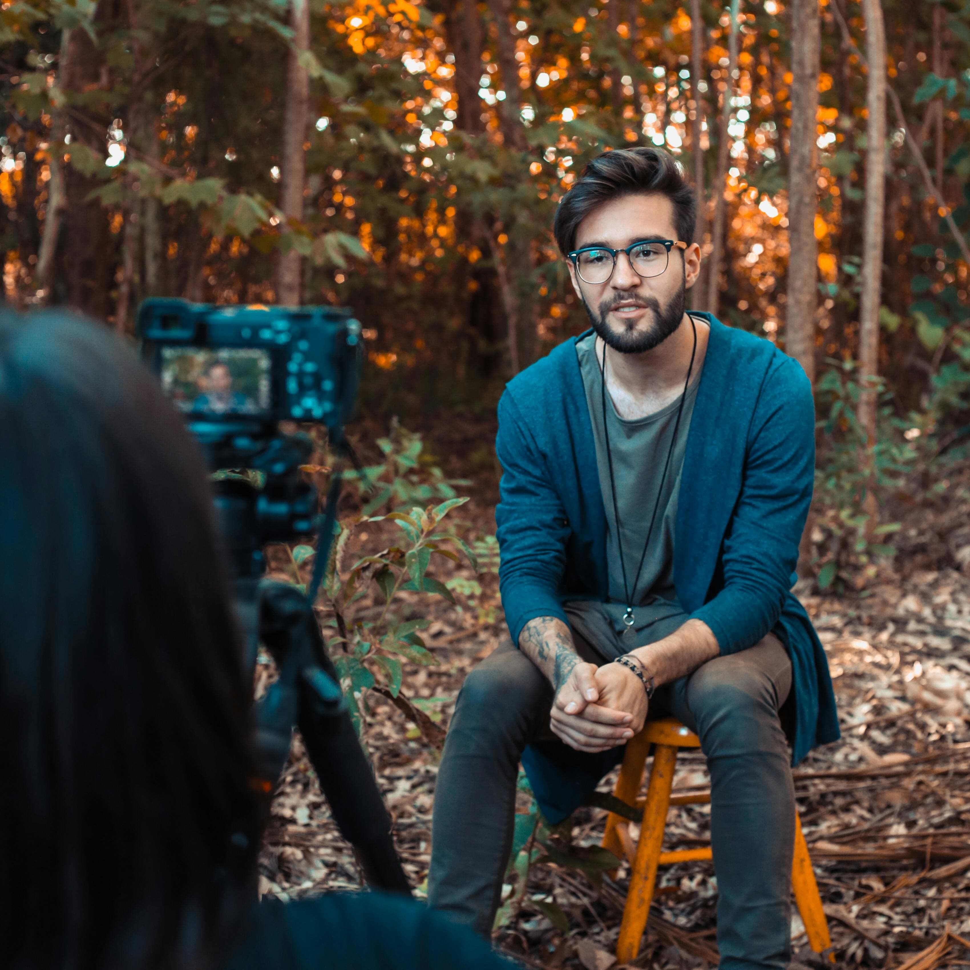 A man with glasses sits on a stool in a forest setting, being filmed for a video. His casual yet professional demeanor represents the importance of authenticity and visual appeal when designing the best product landing pages.