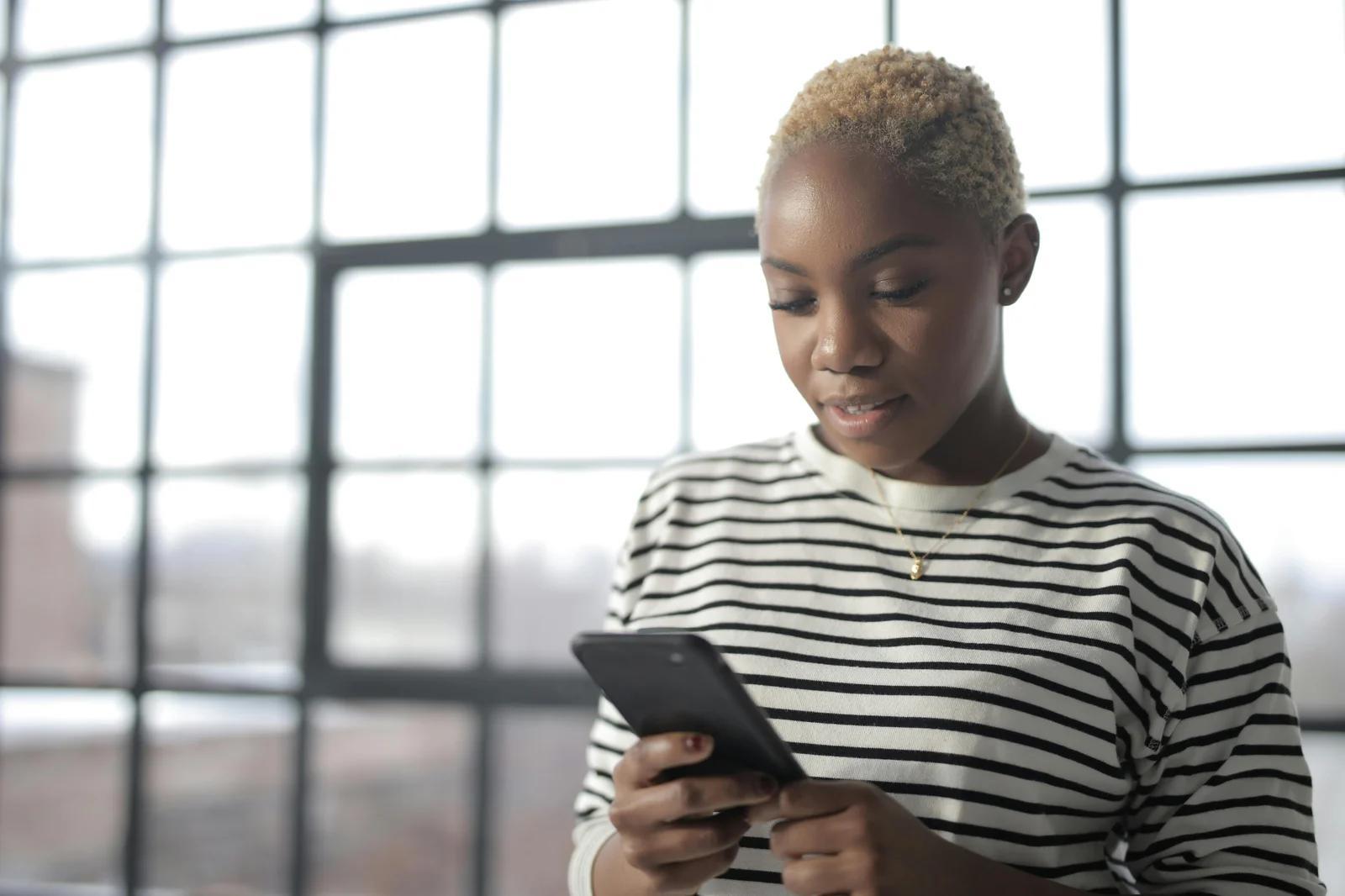 A woman with short blonde hair looking at her phone while standing in front of large windows. She wears a striped sweater. The image portrays a professional working on a video marketing strategy.