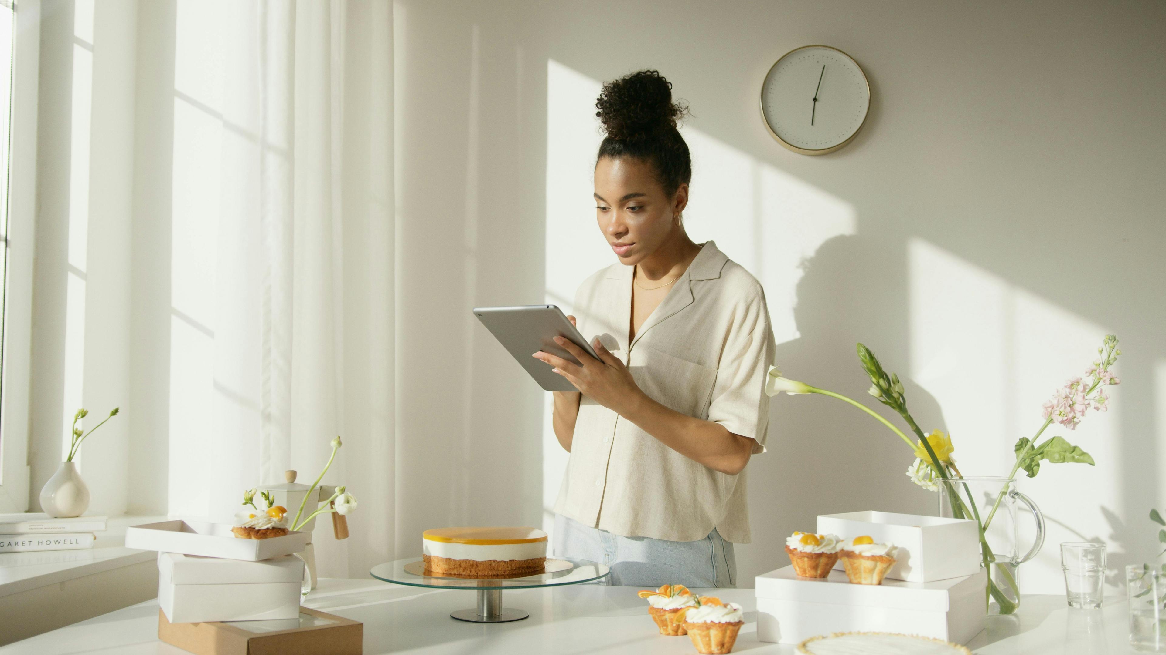 A woman in a bright room with desserts on the table, looking at a tablet, demonstrating engagement with her business. How to market a coaching business.