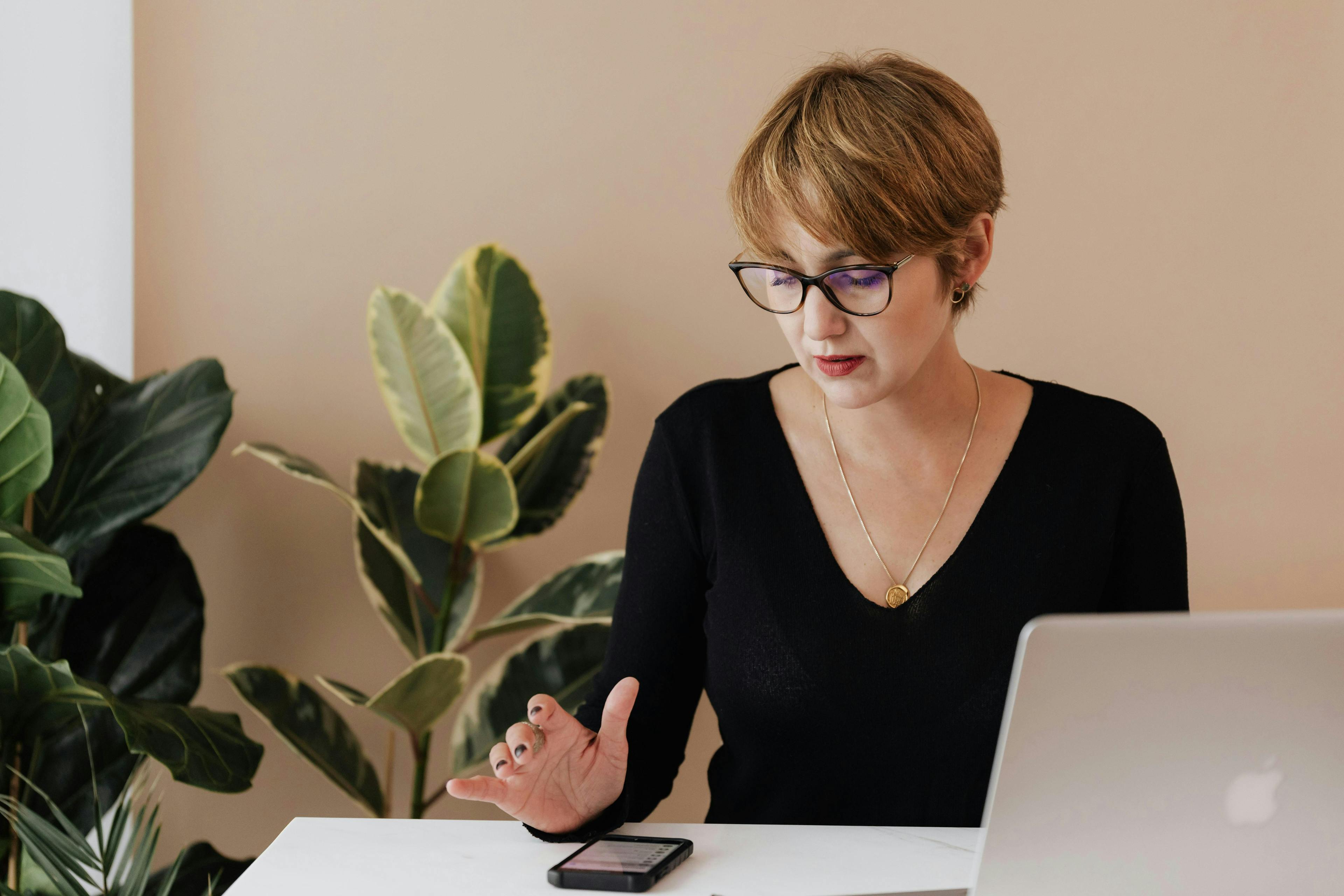 A woman wearing glasses working on her smartphone and laptop, with green plants in the background. This image depicts a marketing consultant for small businesses, highlighting their focus and dedication to client success.