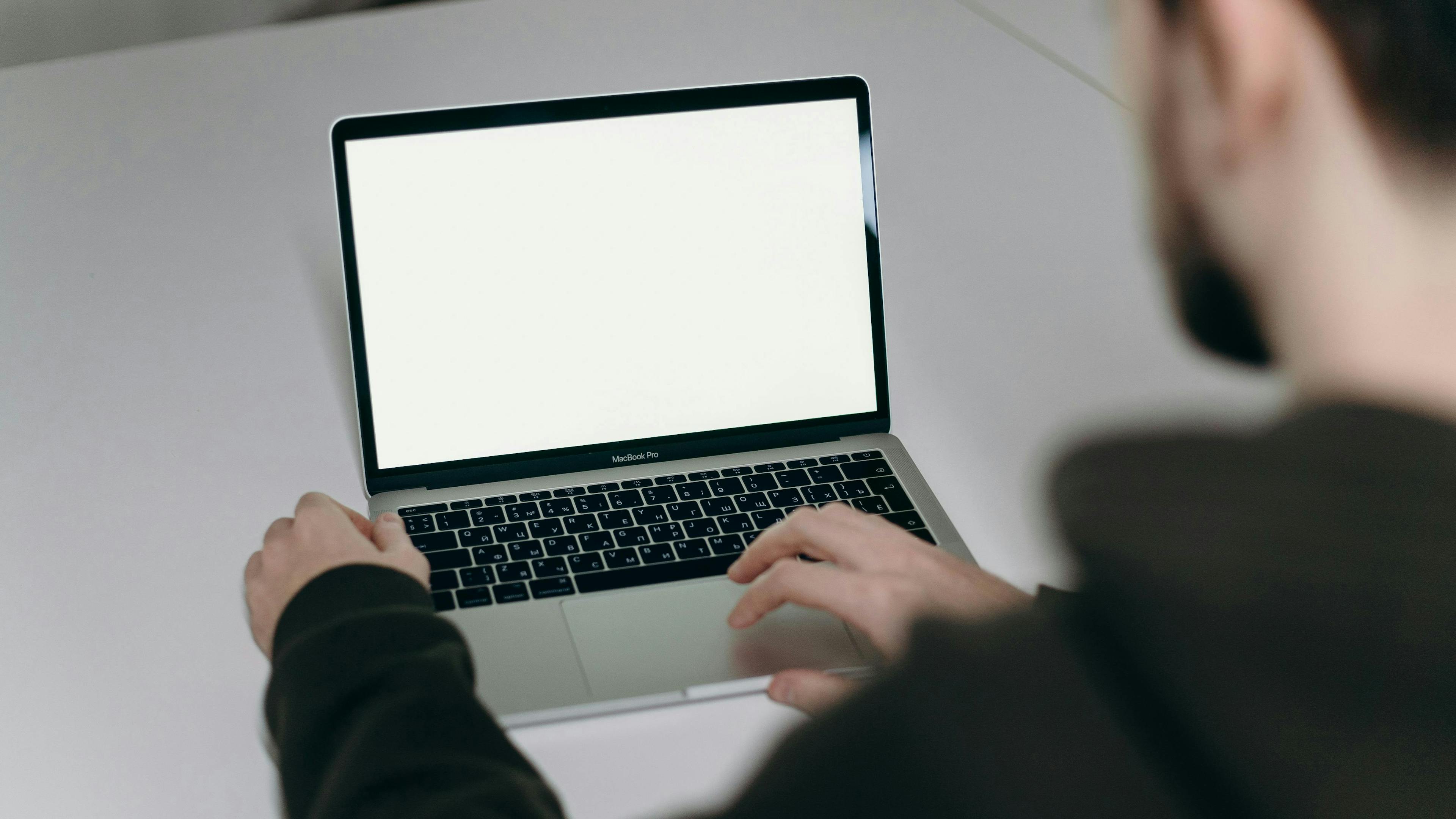 A person with a beard working on a MacBook Pro with a blank screen, sitting at a white desk. The setting is minimalistic, highlighting a focus on digital tasks. The individual appears to be part of an eCommerce marketing agency, possibly setting up a campaign or analyzing data to improve marketing strategies. The clean and simple workspace reflects a professional and organized environment.