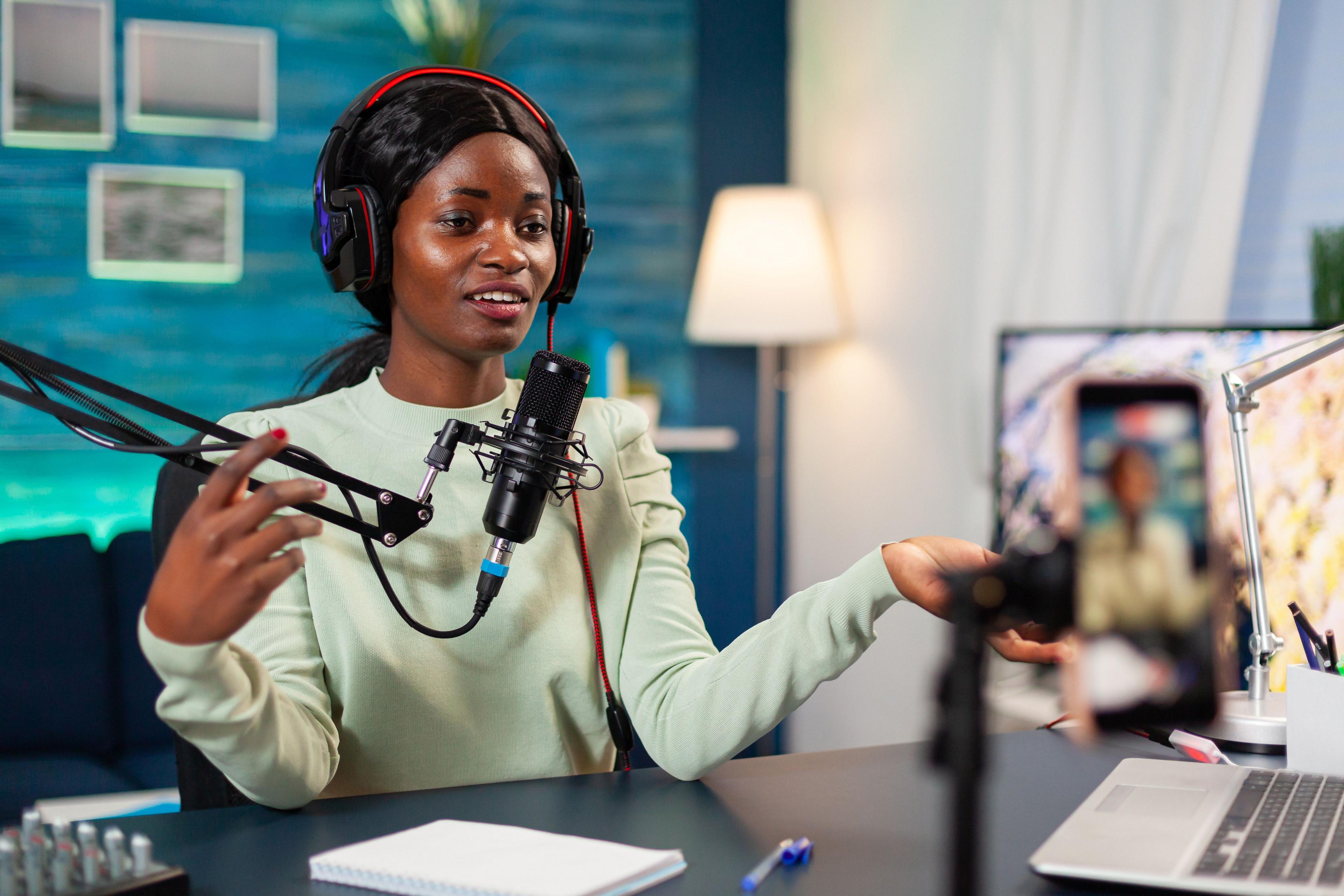 A man and woman in a radio studio setting are engaged in conversation. Both wear headphones and speak into microphones, with plants and cozy decor in the background. 