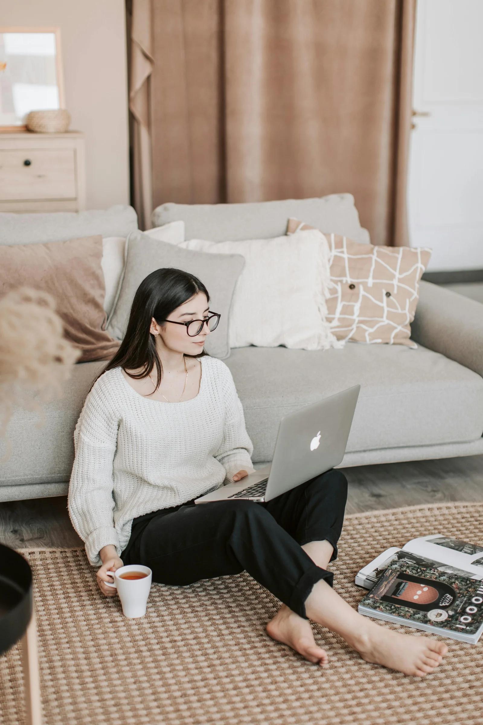 A woman sitting on the floor in a cozy living room, working on a laptop while holding a cup of tea. Several magazines are scattered around her, suggesting she is planning or researching for a video marketing strategy.