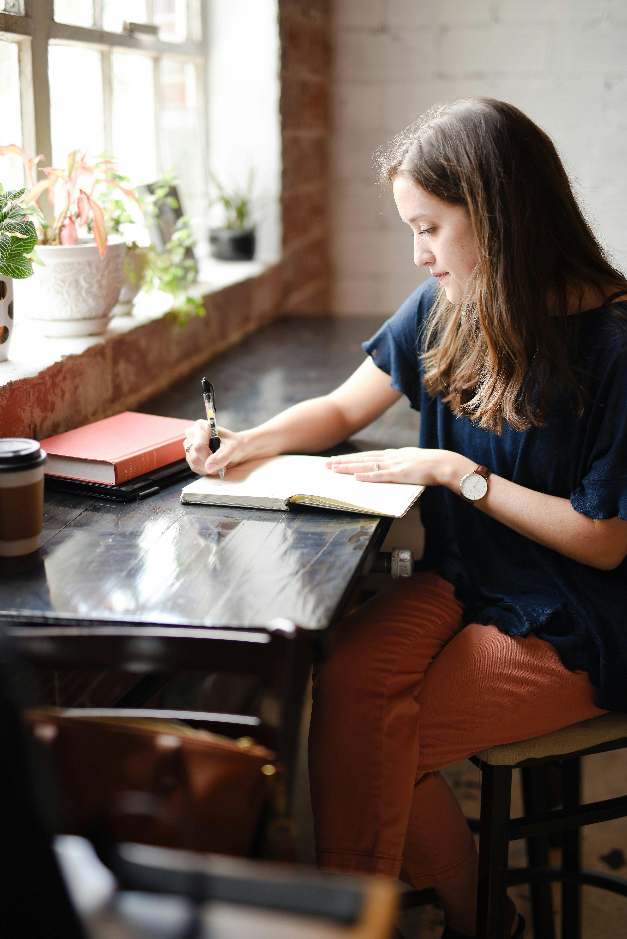 A woman sitting by a window in a café, writing in a notebook with a cup of coffee nearby, possibly outlining steps for a data project involving a train test split in sklearn.