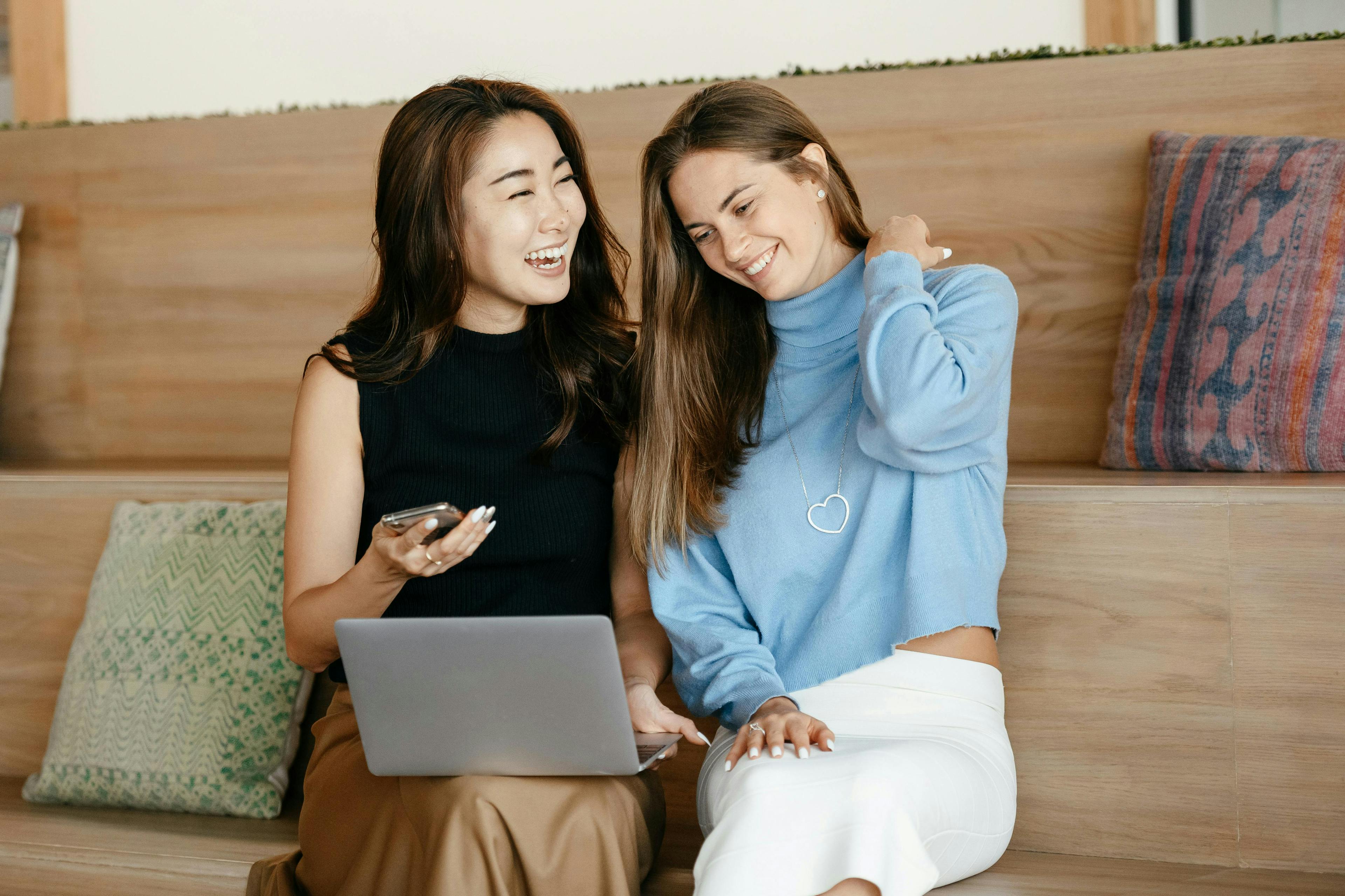 Two women sitting and laughing while working on a laptop and holding a smartphone, representing copywriter AI.