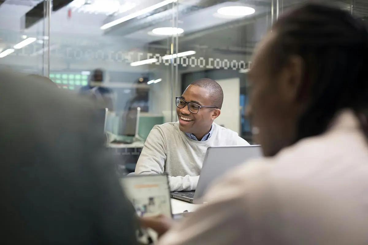 A group of professionals are engaged in a lively discussion in a modern office setting. In the foreground, a person is holding a laptop displaying information. The focus is on a smiling man wearing glasses and a light grey sweater, appearing engaged and enthusiastic. The background shows a glass wall with abstract patterns and other colleagues in conversation. The atmosphere is collaborative and dynamic, embodying the spirit of "audio marketing services."