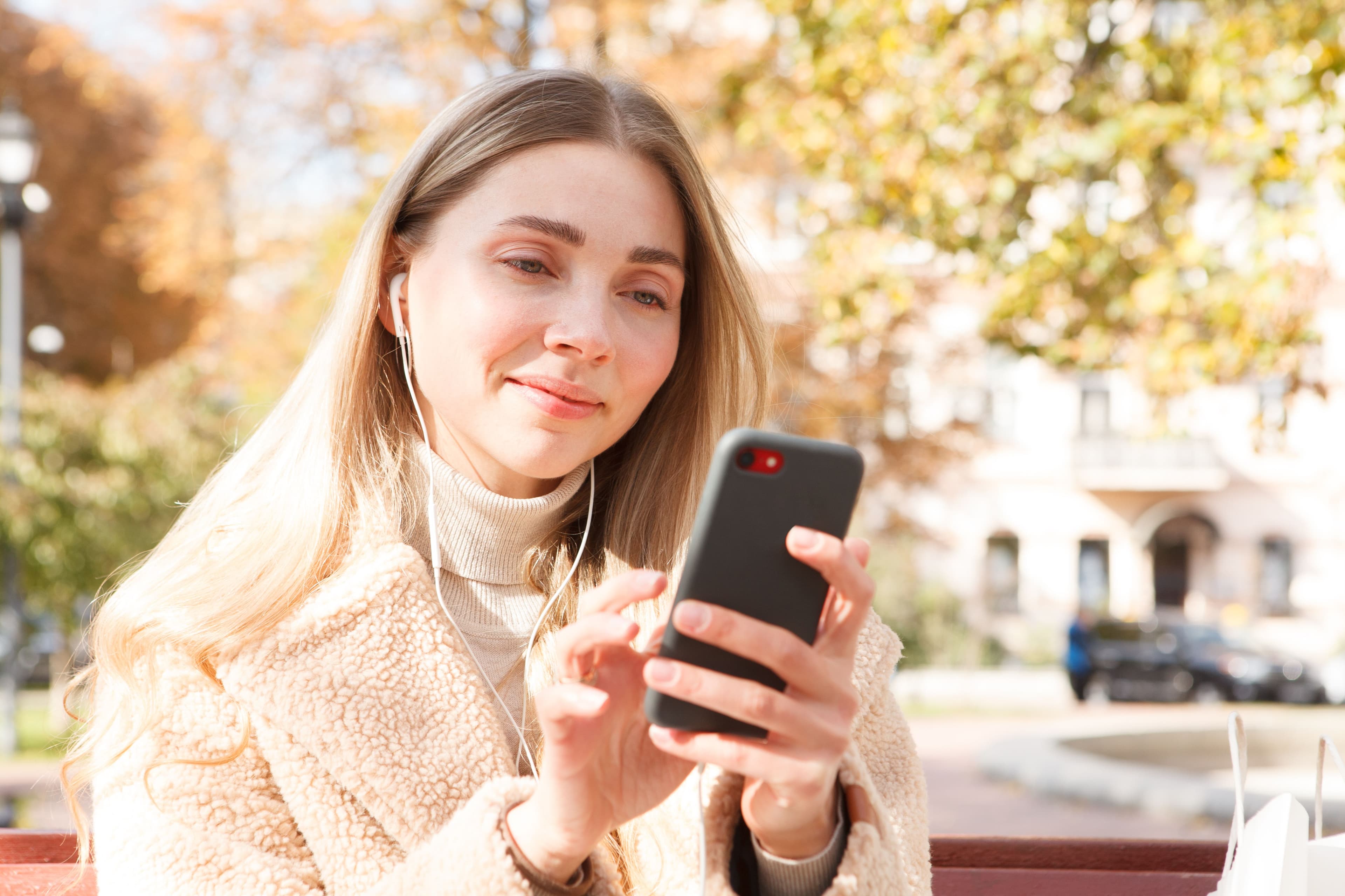 A young woman sitting on a park bench, smiling while scrolling through her smartphone, symbolizing social media engagement and mobile marketing.